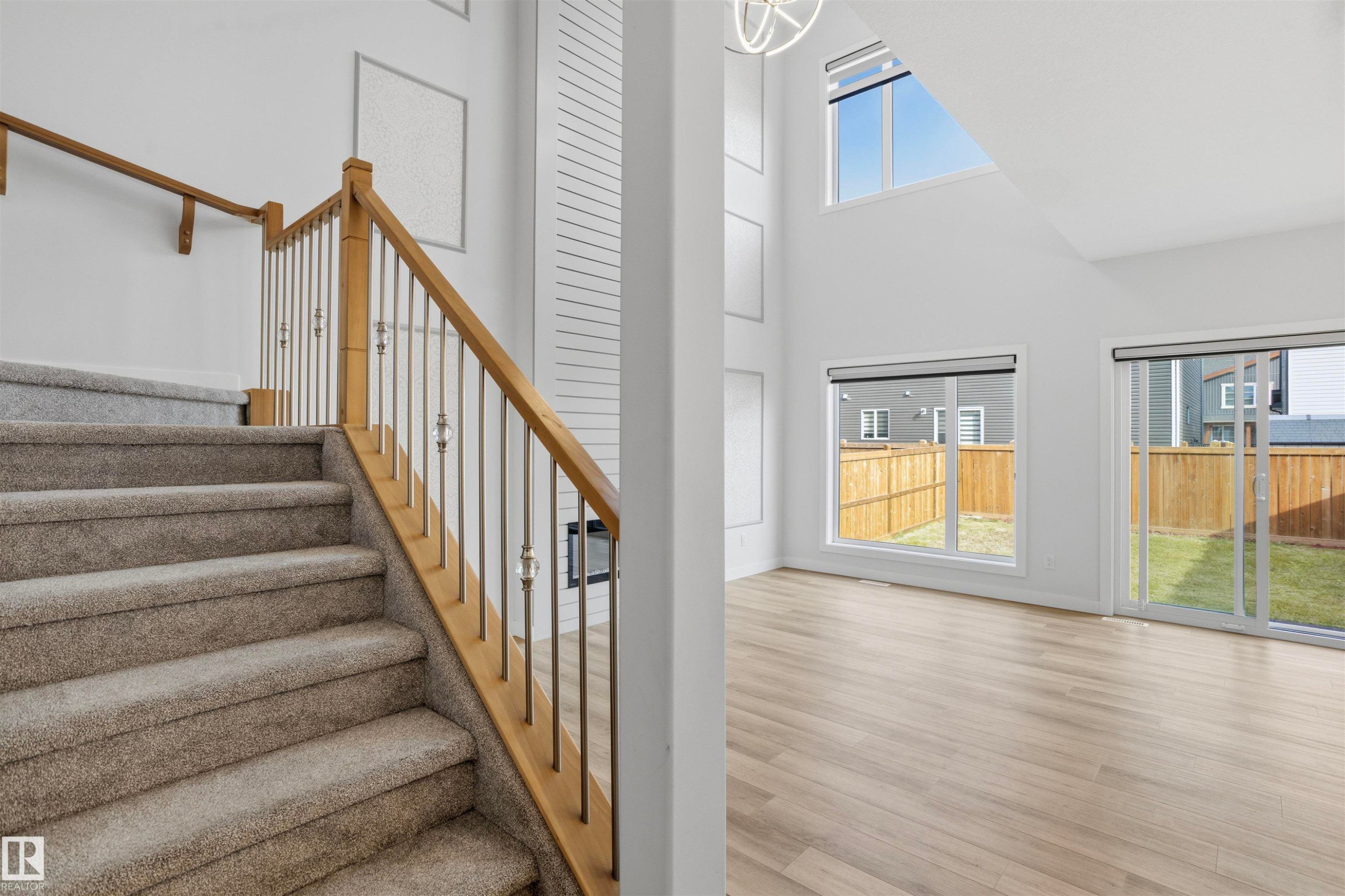 Double-height living area featuring wood-finish flooring, floor-to-ceiling fireplace surround with horizontal detailing, staircase with wooden handrail, and sliding glass doors - 2045 209A St Nw, Edmonton, AB