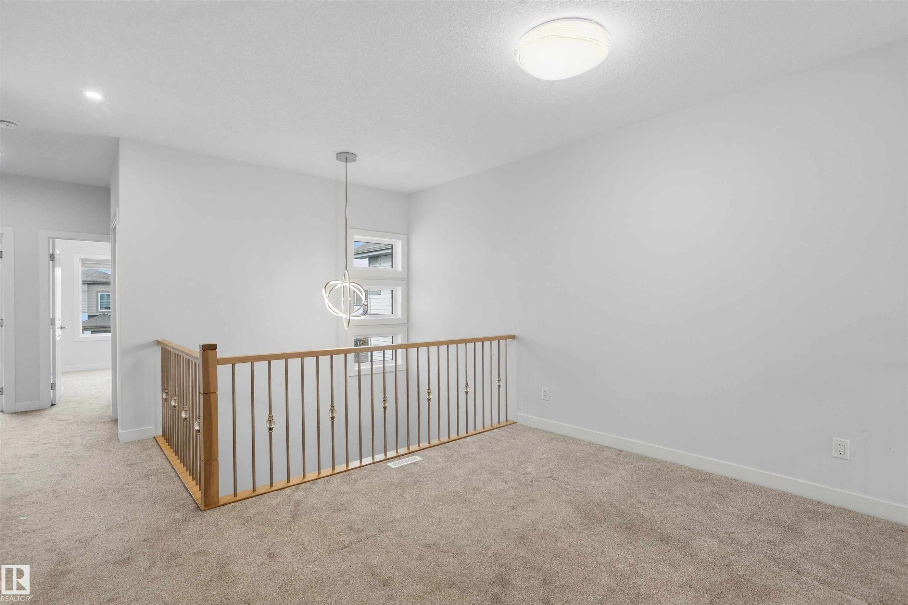 Open floor space featuring neutral carpeting, a staircase with wood handrails and metal balusters, stacked windows, and a multi-ring spherical pendant light - 2045 209A St Nw, Edmonton, AB