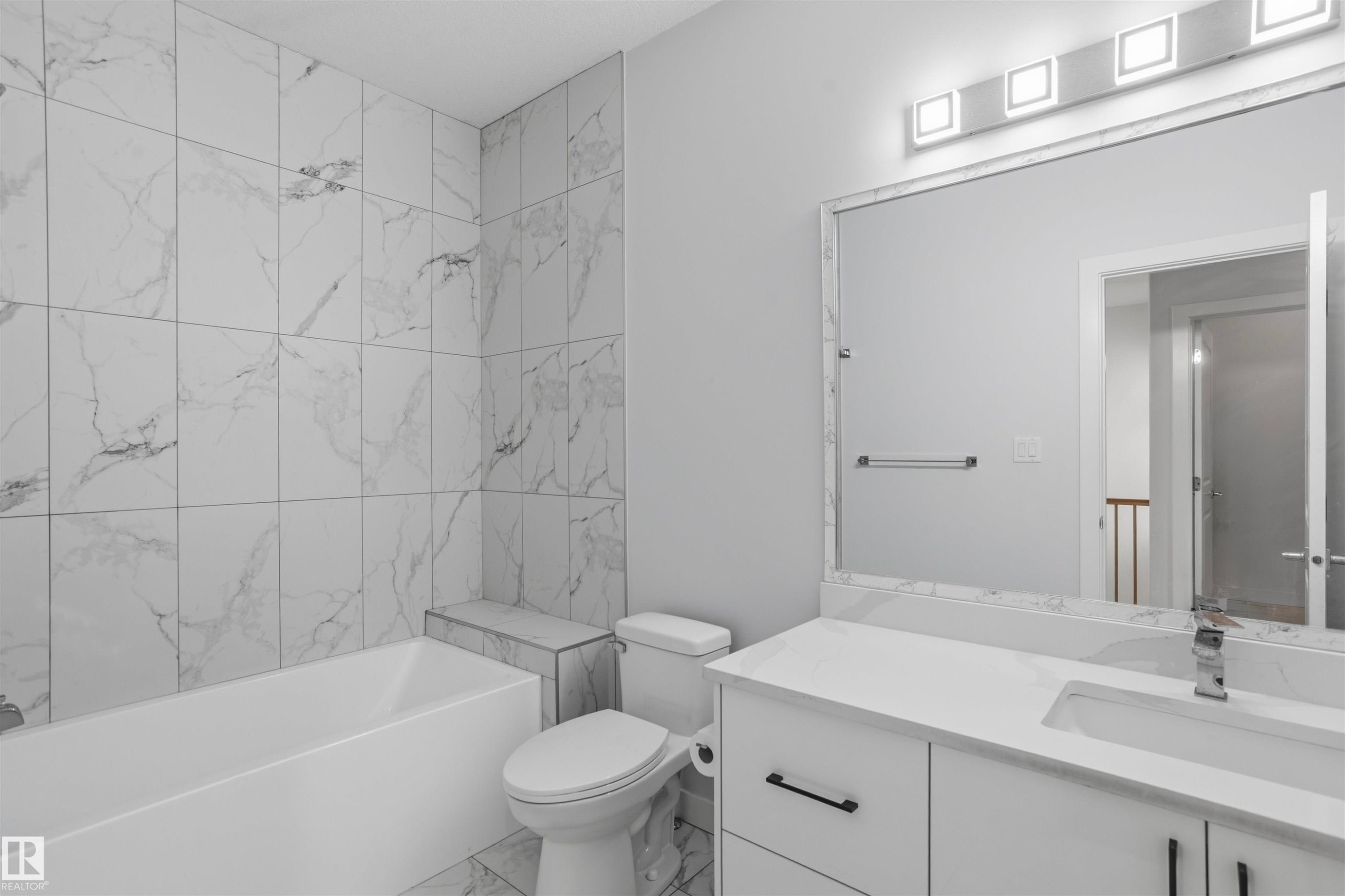 Full-height shower surround with large-format stone-patterned tile, white vanity with integrated basin and quartz countertop, rectangular mirror with tile border, and four-light vanity fixture - 2045 209A St Nw, Edmonton, AB