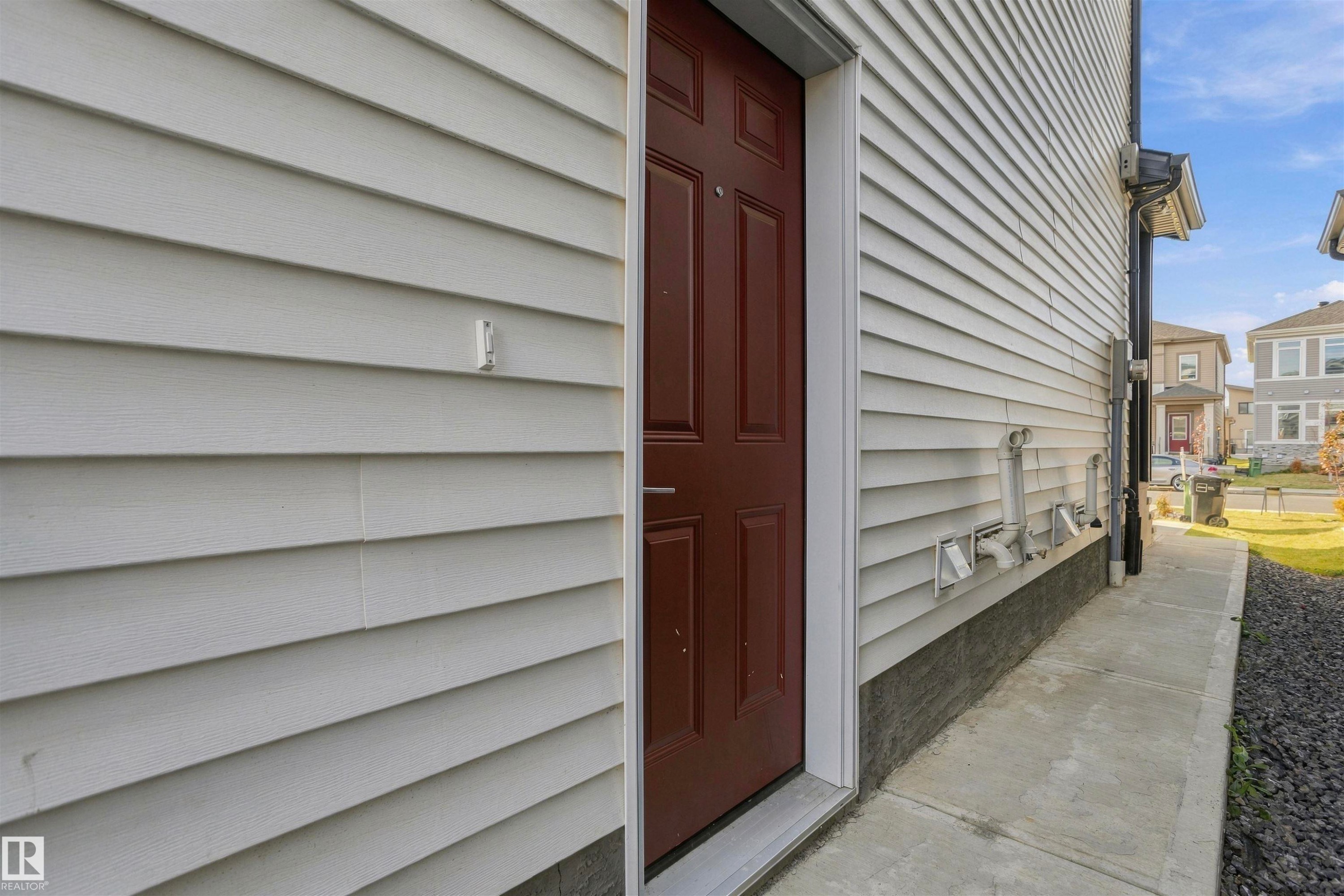 Exterior siding in a light-toned finish featuring a six-panel deep-red door, concrete walkway, and stone-grounded foundation - 2045 209A St Nw, Edmonton, AB