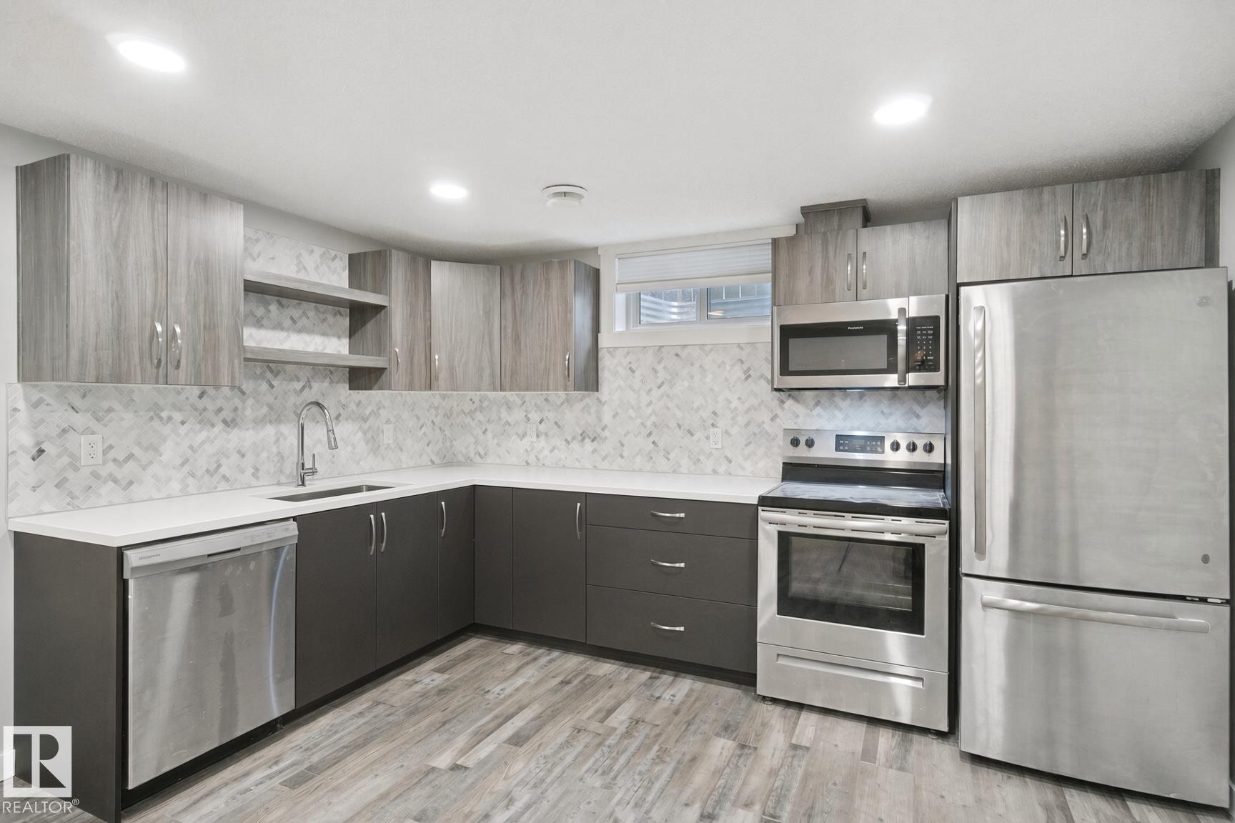 Modern kitchen featuring stainless steel appliances, a herringbone tile backsplash, gray base cabinetry, wood-finish upper cabinets, and light-toned countertops - 13212 73 Street Nw, Edmonton, AB