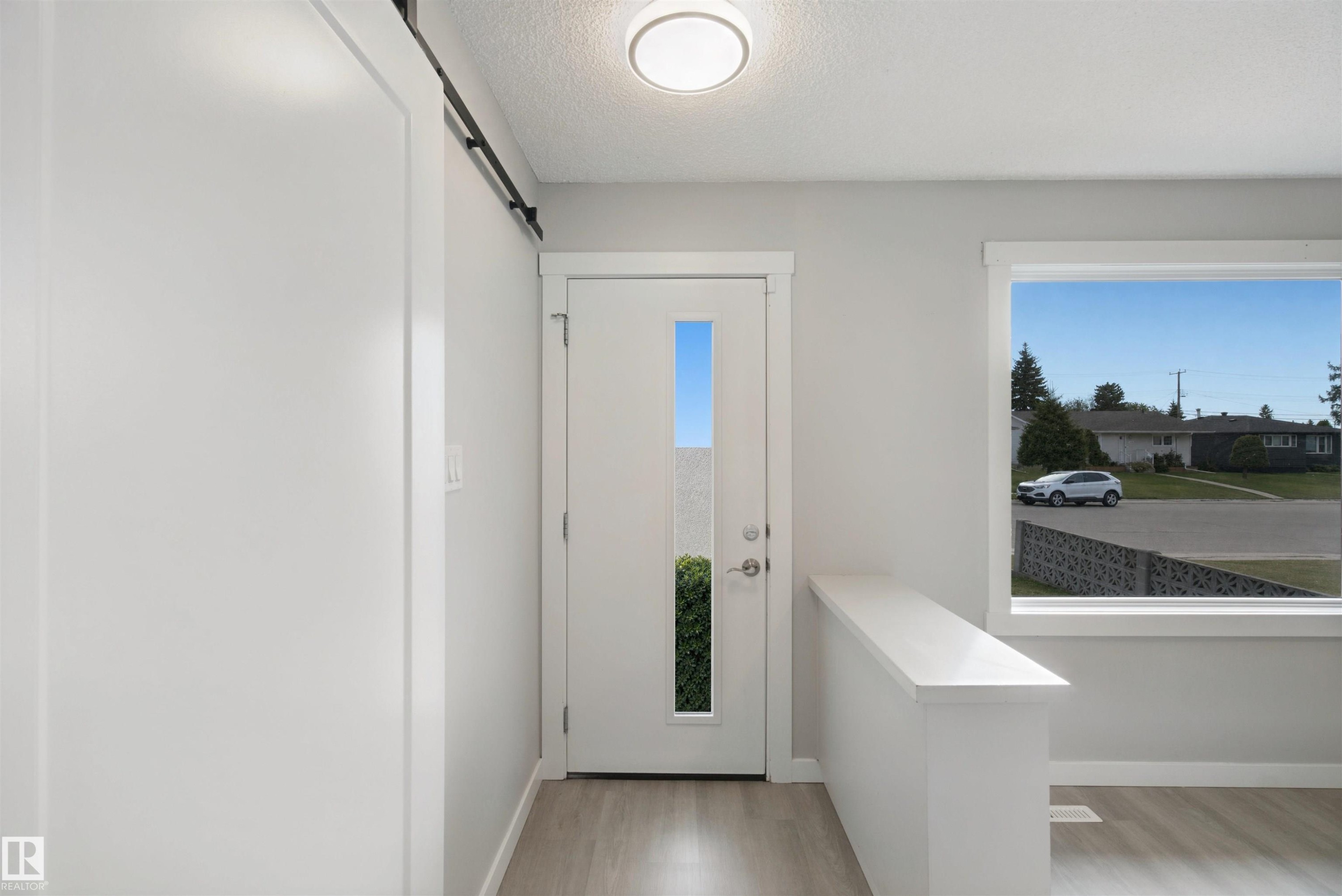 Contemporary entryway featuring a front door with vertical window insert, a side-mounted barn door track, wood-finish flooring, and a large picture window - 13212 73 Street Nw, Edmonton, AB