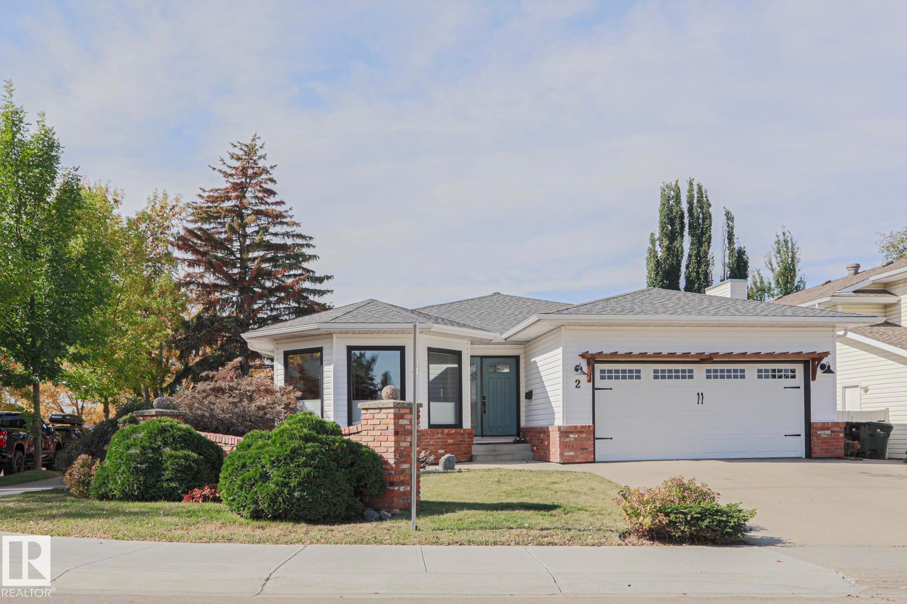 White siding exterior with brick accents, black-framed windows, an attached two-car garage, and a landscaped front yard with mature shrubbery - 2 Hillview Crescent, Sherwood Park, AB