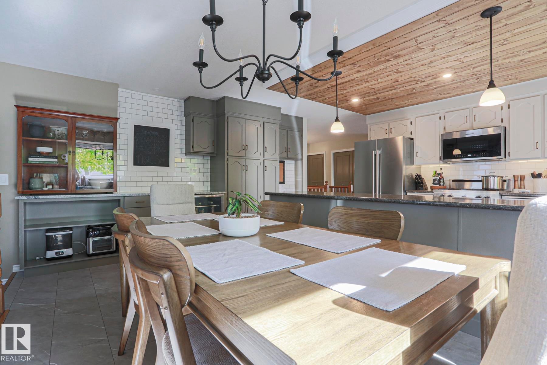 Integrated dining area with chandelier lighting and wood-finish ceiling, featuring a kitchen with stainless steel appliances, subway tile backsplash, and dark countertops - 2 Hillview Crescent, Sherwood Park, AB