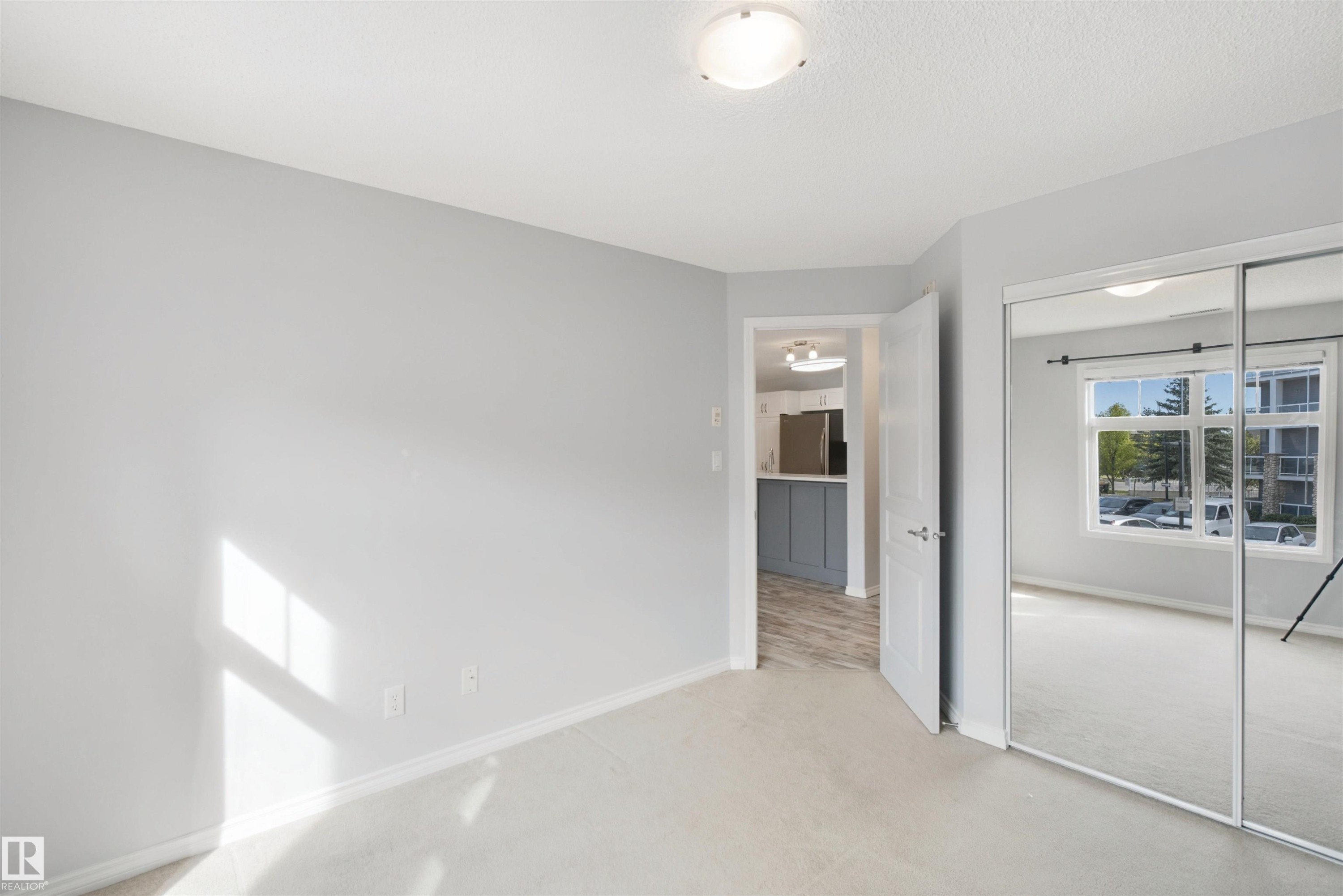 Neutral-toned walls, carpet flooring, mirrored sliding closet doors, and a flush mount ceiling light fixture - #208 16035 132 St Nw, Edmonton, AB