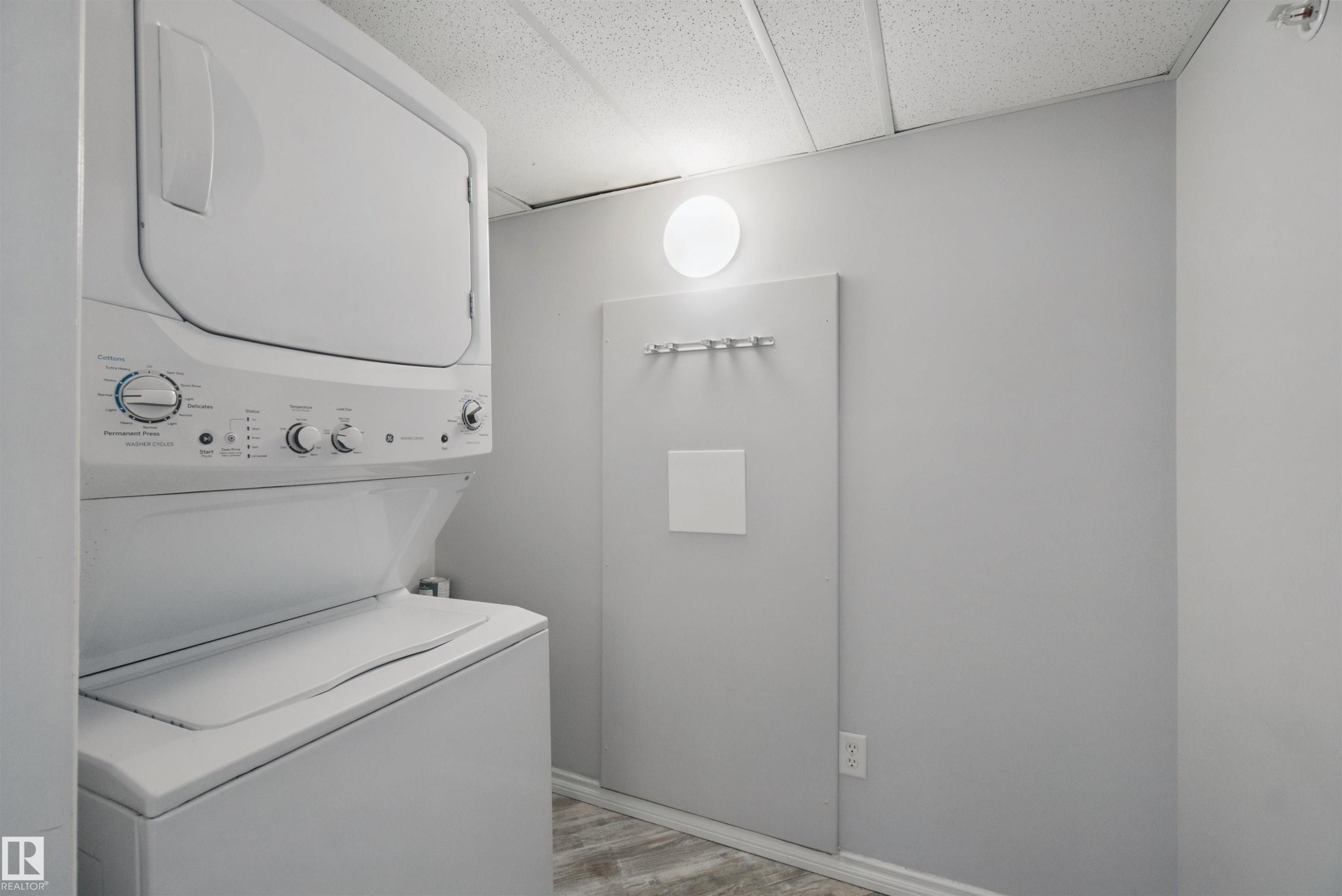 Stackable washer and dryer, light gray wall color, wood-finish flooring, and round wall-mounted light fixture - #208 16035 132 St Nw, Edmonton, AB