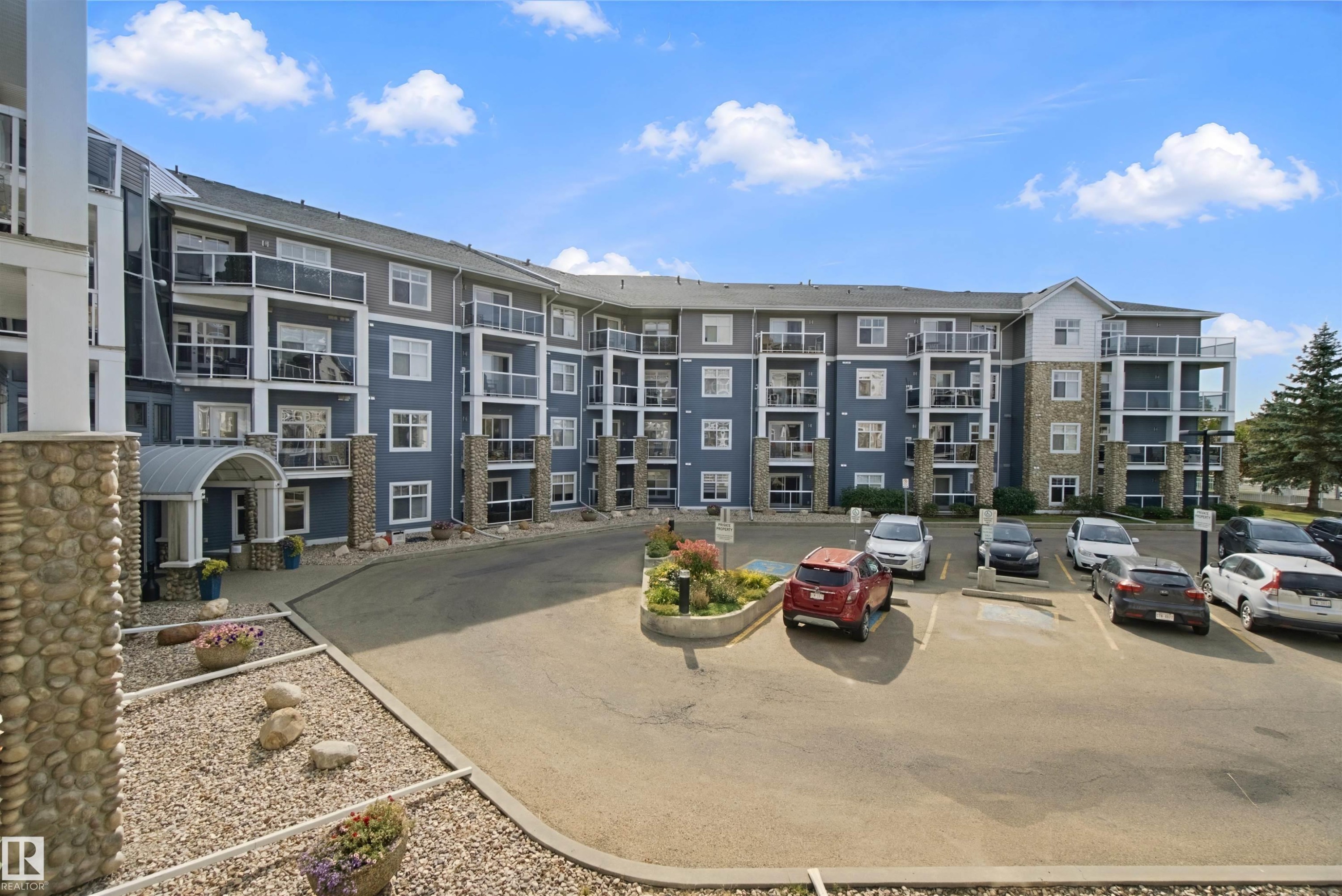Multi-story residential building with stone and siding exterior, multiple private balconies with glass railings, covered main entrance, and an on-site parking area - #208 16035 132 St Nw, Edmonton, AB