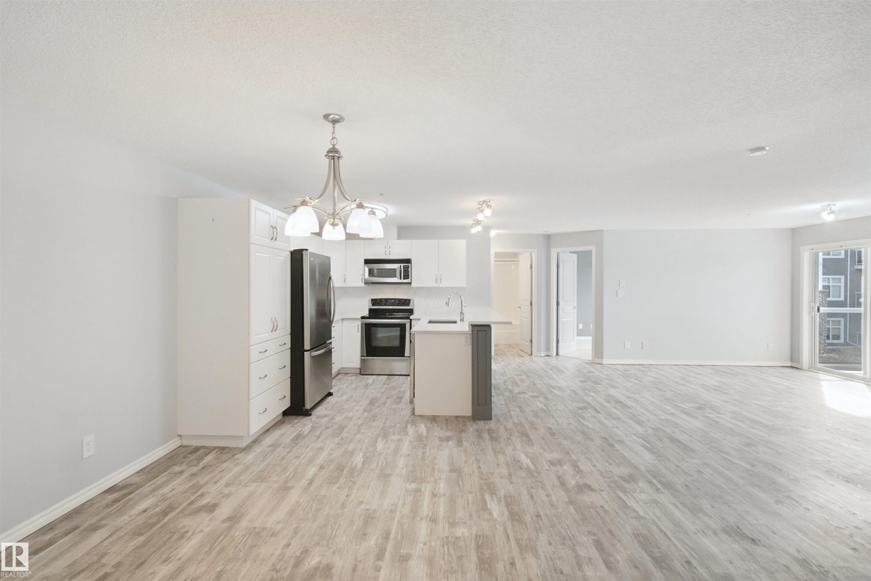 Bright open concept living area featuring wood-finish flooring, white cabinetry, stainless steel appliances, and a sliding glass patio door - #208 16035 132 St Nw, Edmonton, AB