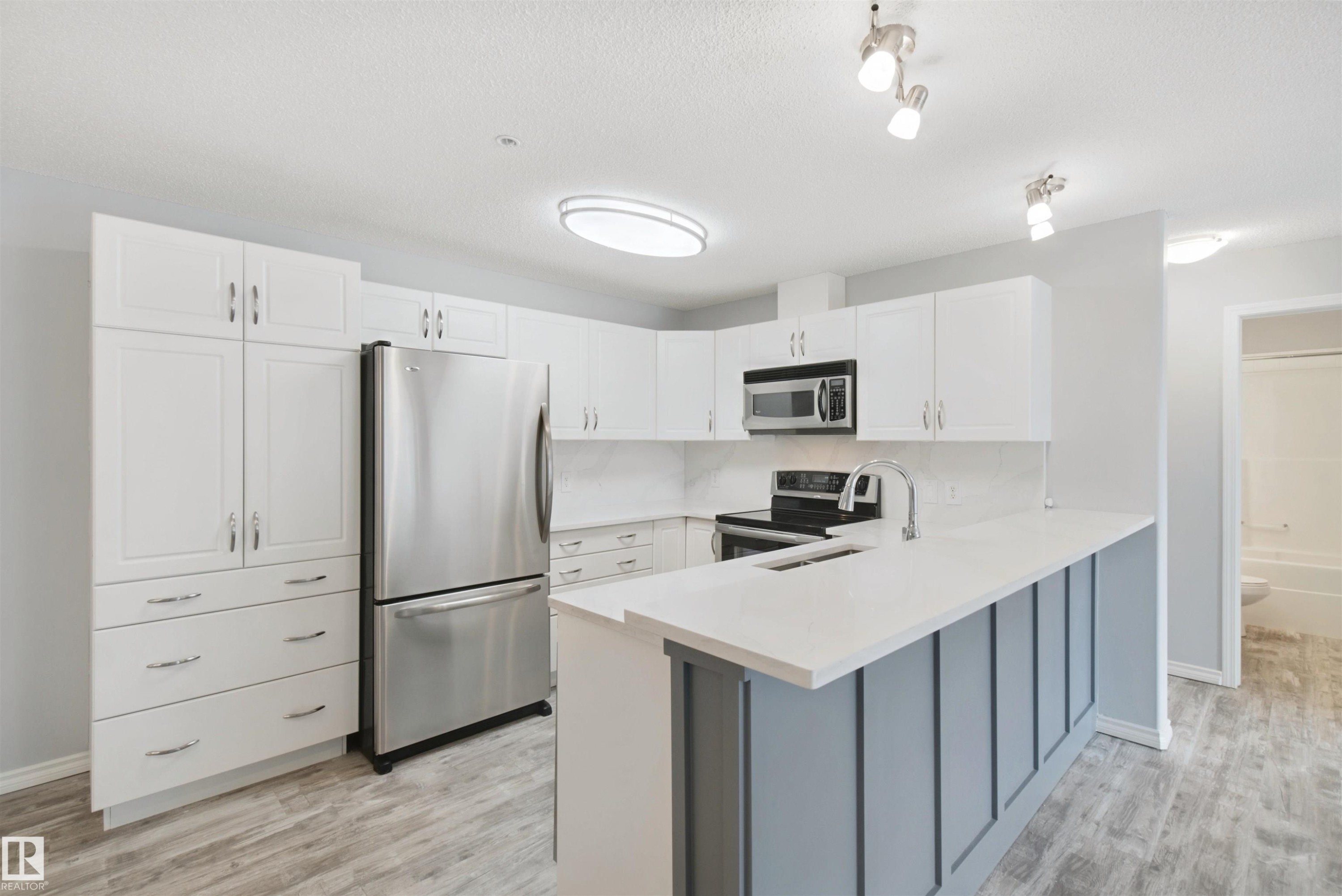 Kitchen featuring white cabinetry, stainless steel appliances, a white countertop, wood-finish flooring, and a breakfast bar - #208 16035 132 St Nw, Edmonton, AB