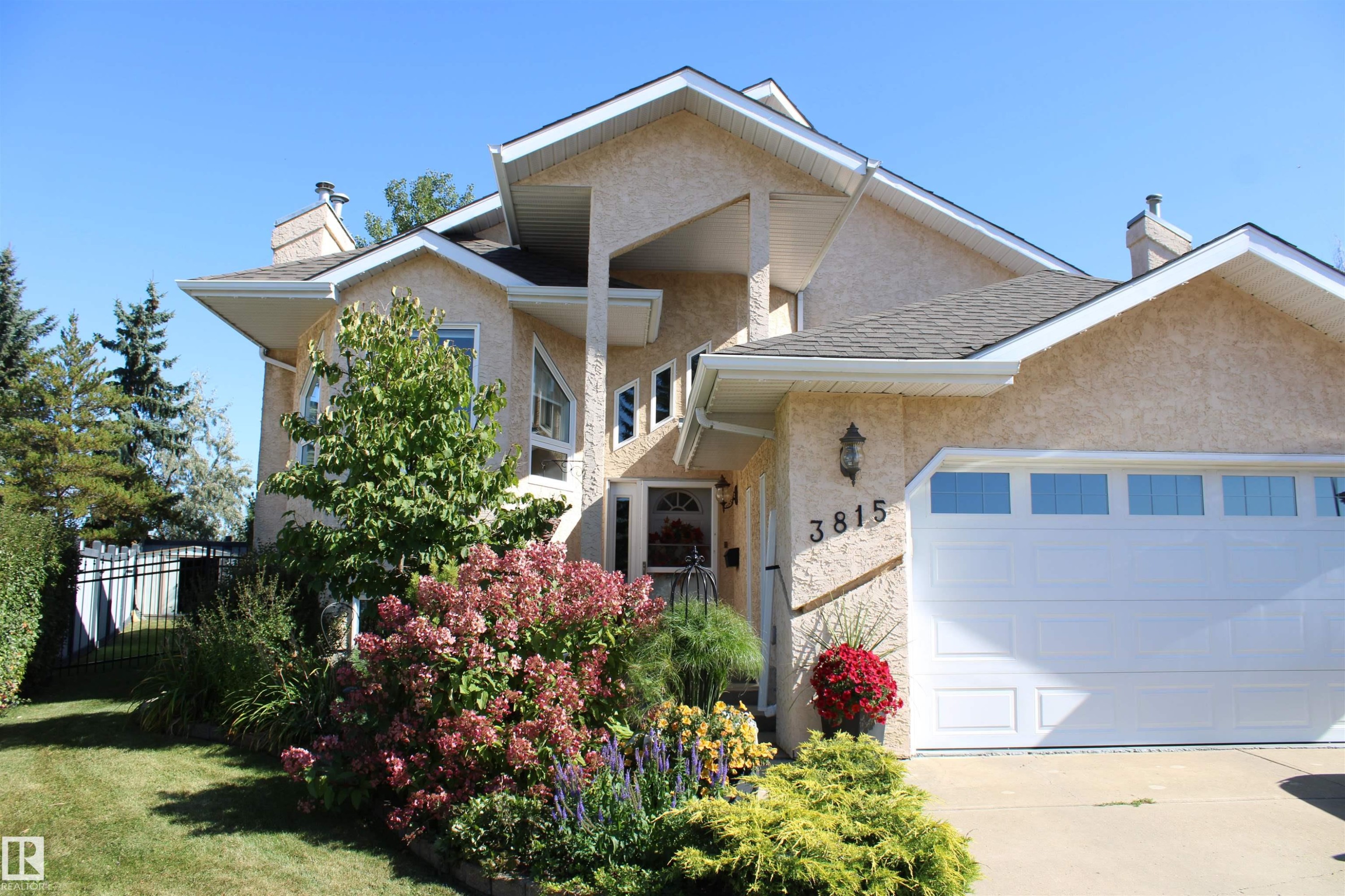 Stucco exterior with a peaked roofline, an attached heated two-car garage with transom windows, a front entry with a decorative light fixture, and established front landscaping - 3815 41 Street Nw, Edmonton, AB