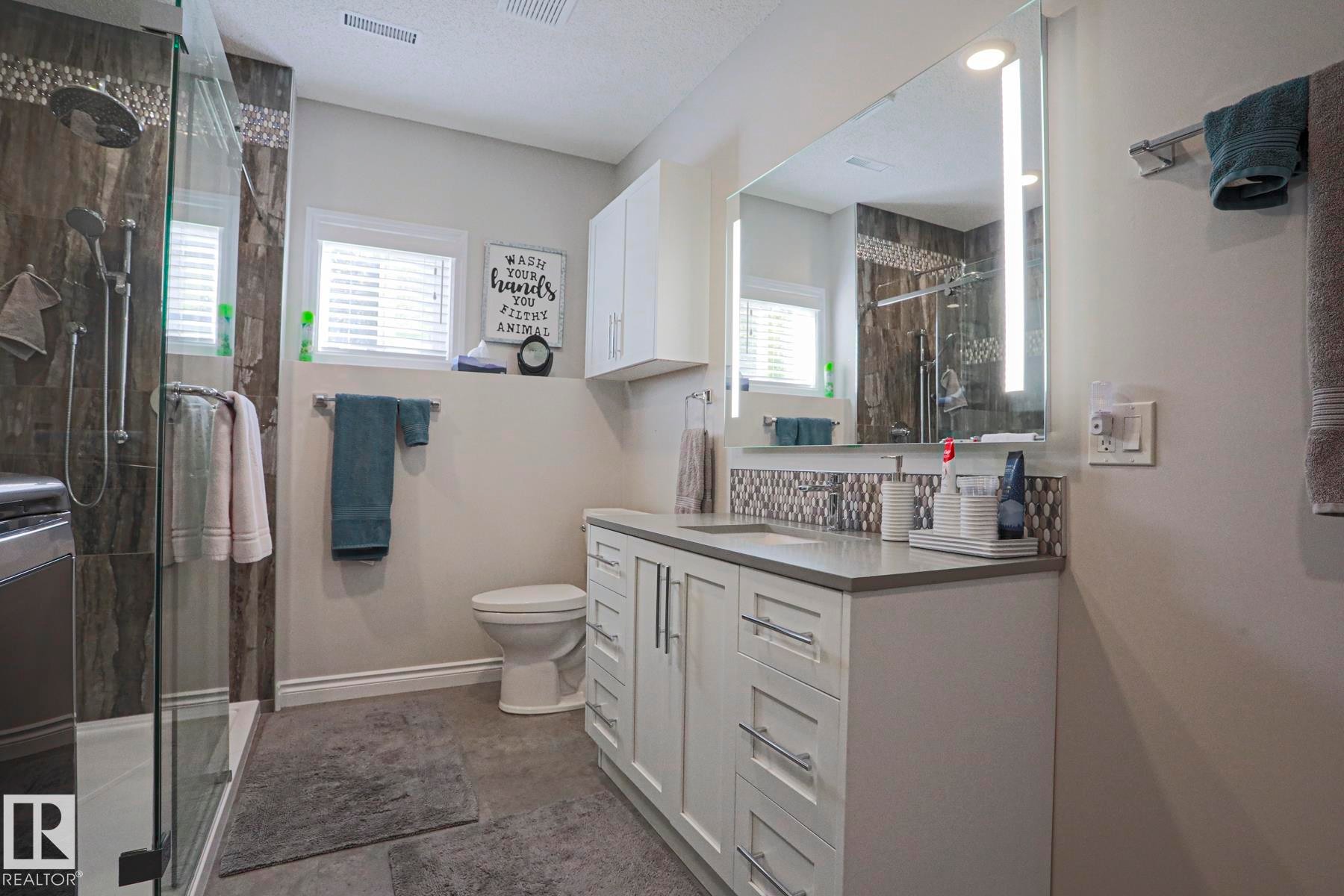 Walk-in shower with glass enclosure and rain showerhead, vanity with undermount sink and stone-finish countertop, illuminated mirror, and mosaic tile backsplash - 3815 41 Street Nw, Edmonton, AB