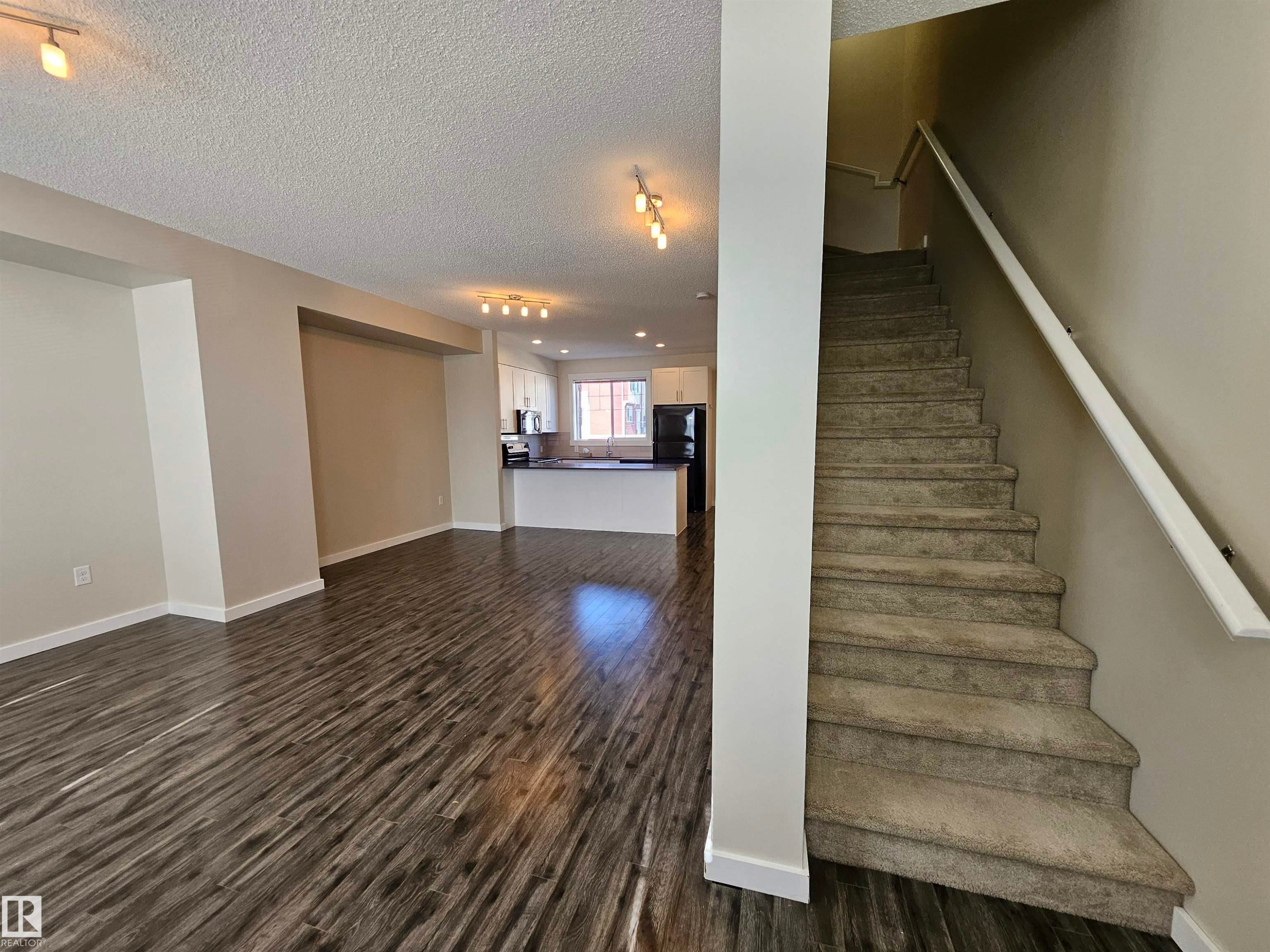 Wood-finish flooring, recessed lighting, white kitchen cabinetry, kitchen island with dark countertop, and carpeted staircase - #69 903 Crystallina Nera Wy Nw, Edmonton, AB