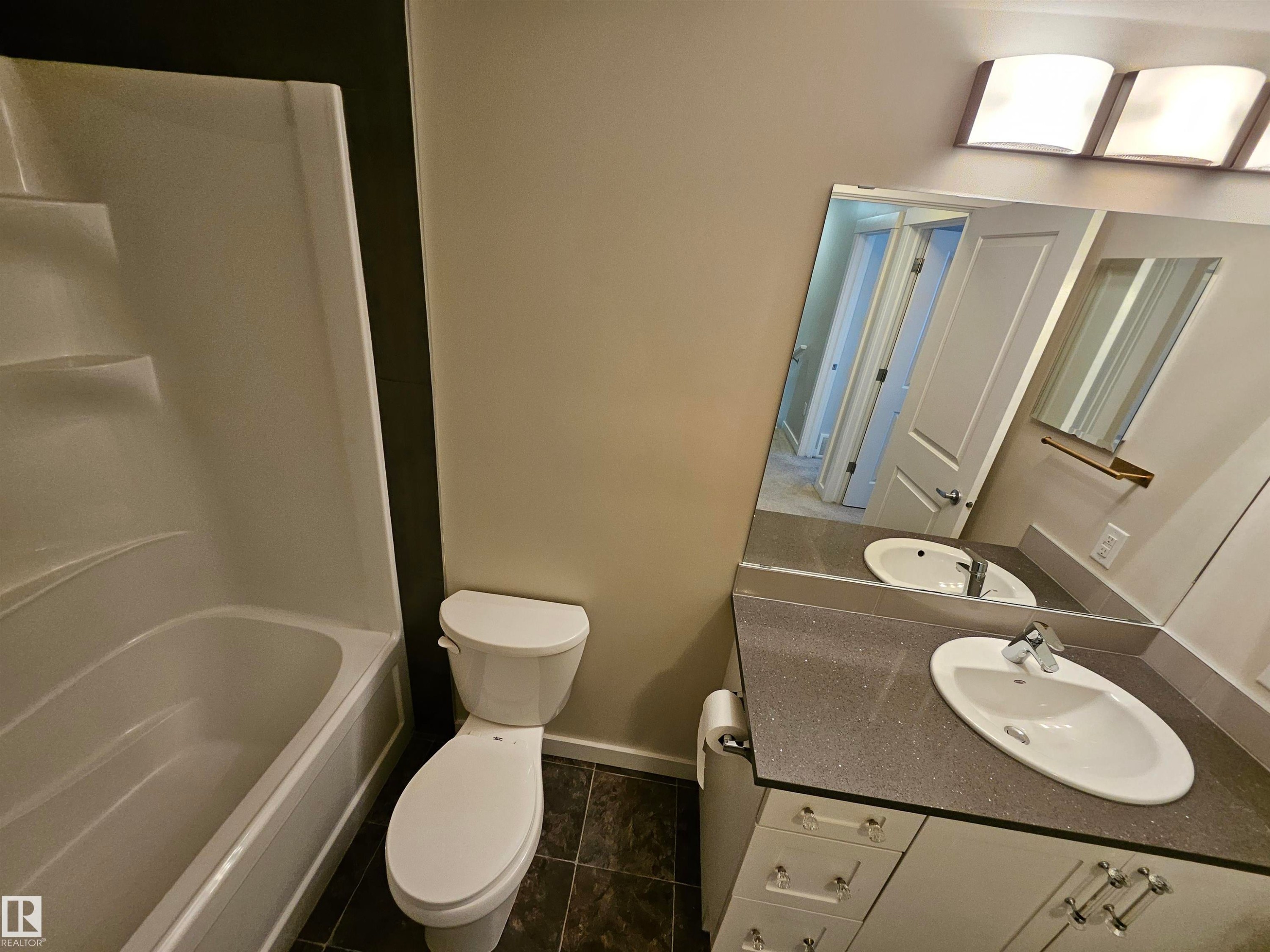 White bathtub with integrated steps, white toilet, dark-toned floor tiling, gray countertop, and a vanity with integrated sink - #69 903 Crystallina Nera Wy Nw, Edmonton, AB