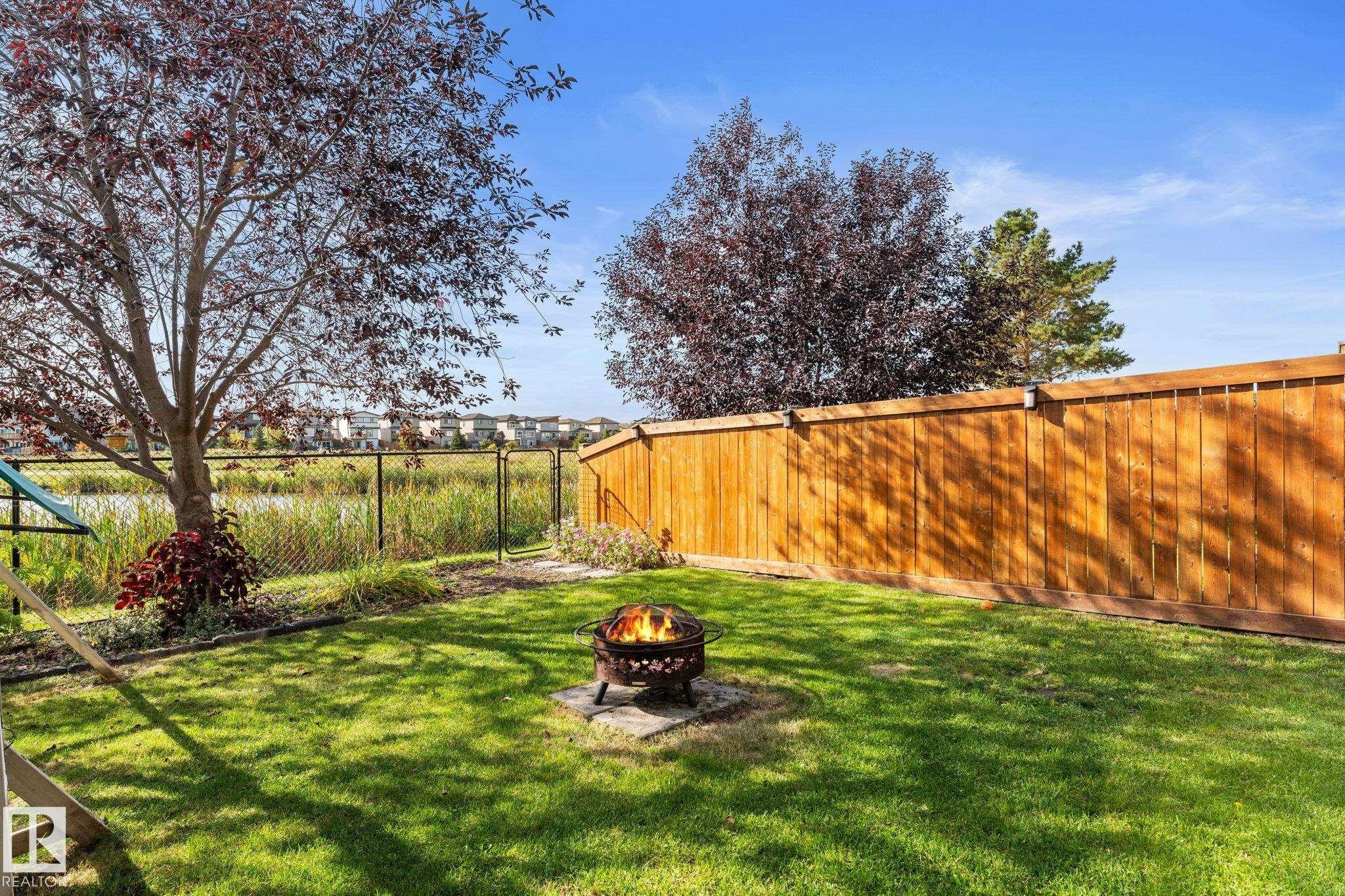 Grassed backyard featuring a wood fence, mature trees, and a chain-link fence bordering a pond - #5 4900 62 St, Beaumont, AB
