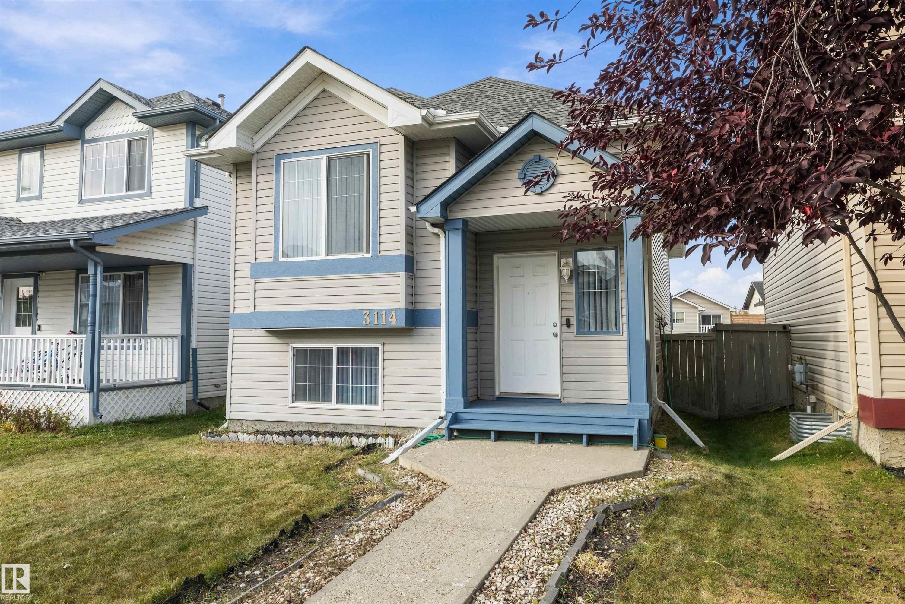 Siding exterior with light blue trim, covered porch entry, mature front yard tree, concrete walkway, and partial fencing - 3114 32 Avenue Nw, Edmonton, AB