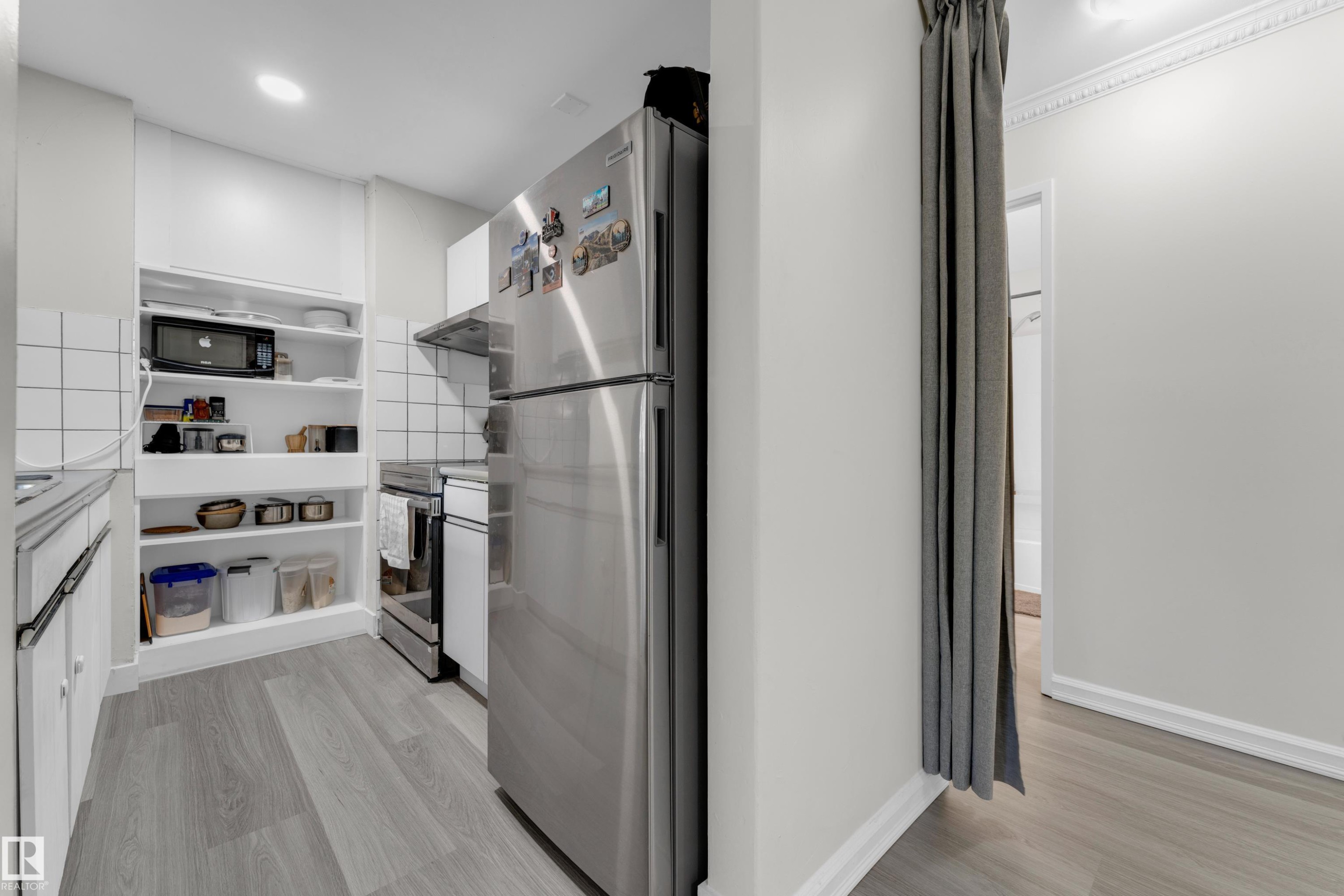 Galley kitchen featuring wood-finish flooring, white subway tile backsplash, stainless steel refrigerator, and integrated open shelving - #113 2508 50 St Nw, Edmonton, AB