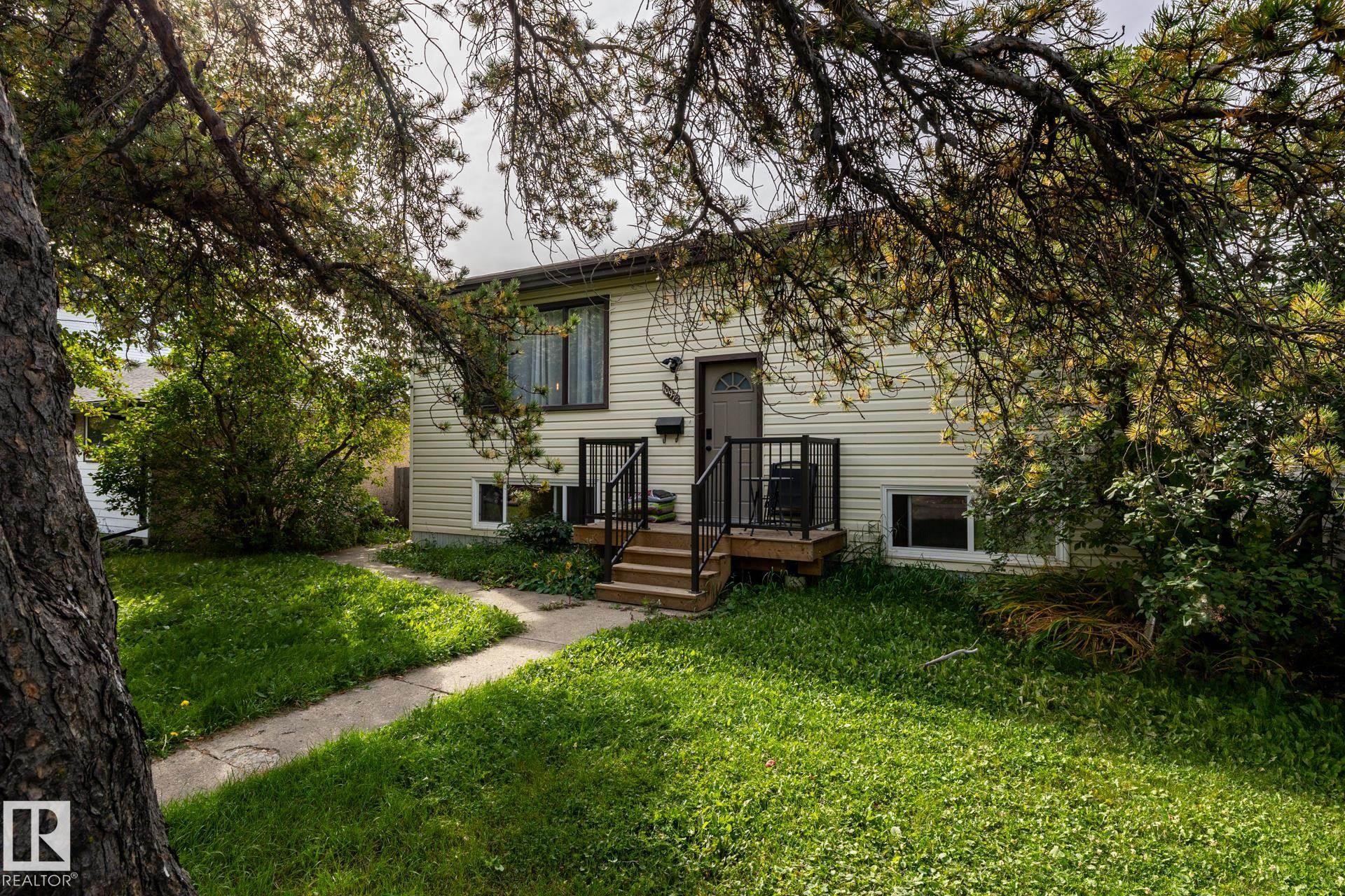Exterior featuring light-colored siding, a front entry with a storm door, an elevated wood-finish deck with black railings, and a concrete pathway - 10972 157 Street Nw, Edmonton, AB