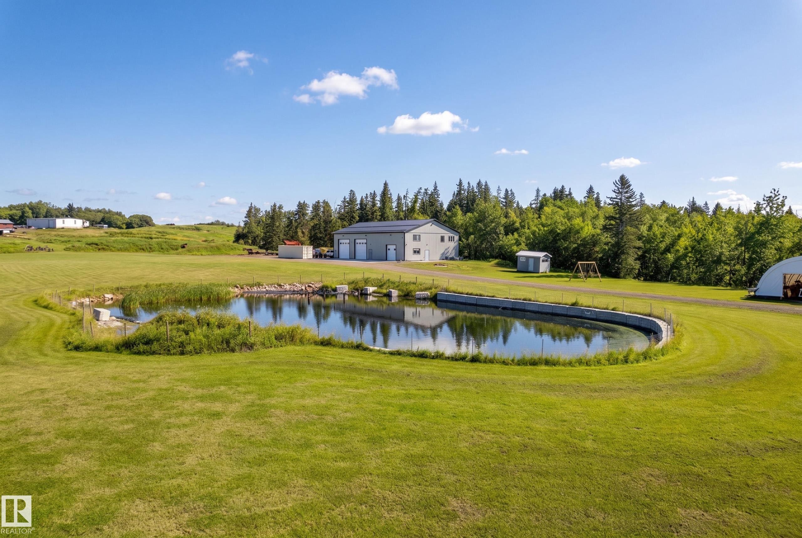 Expansive green space featuring a pond with a concrete retaining wall, a detached multi-bay structure, and a secondary outbuilding - 49279 Rge Rd 240, Rural Leduc County, AB