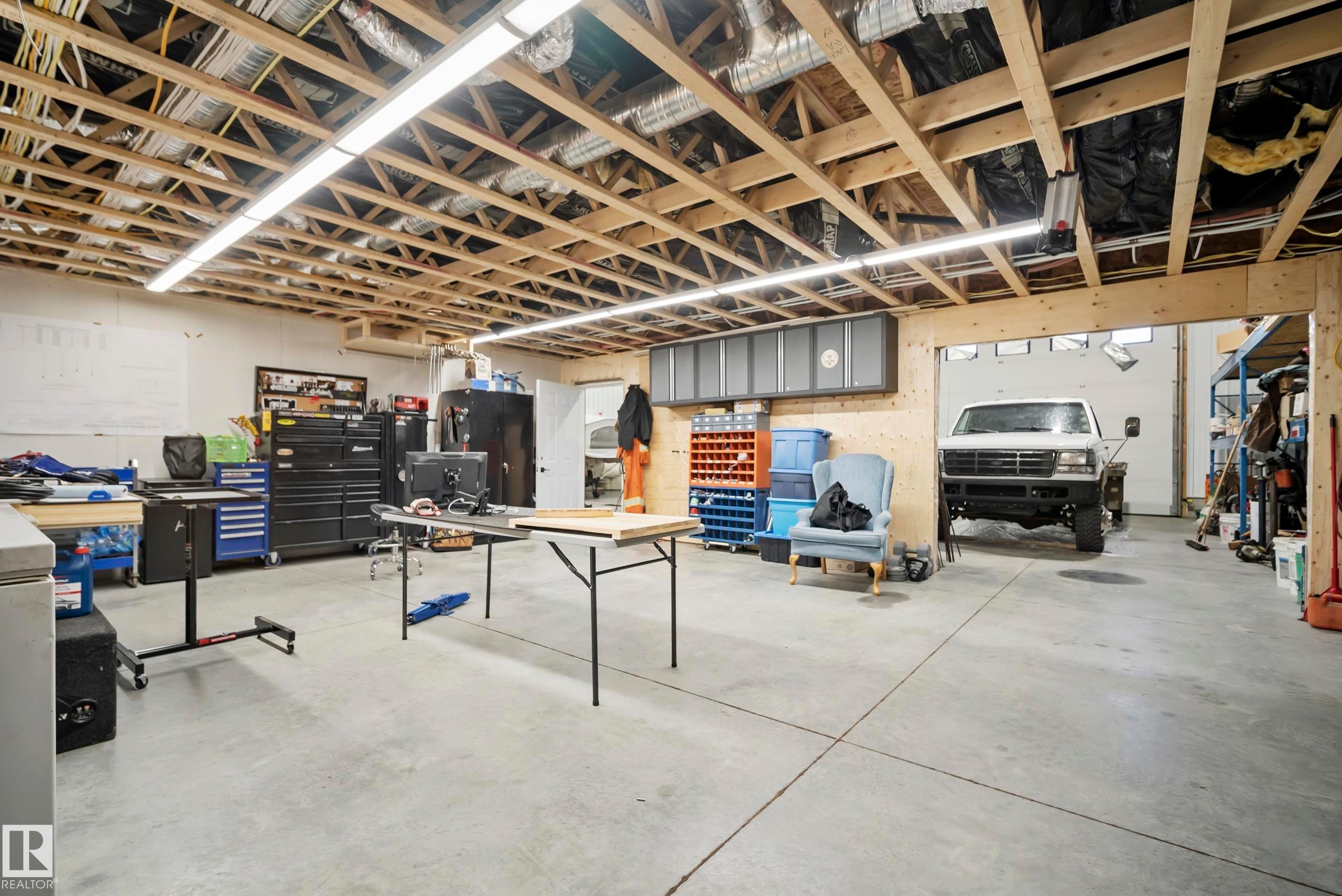Expansive concrete flooring with exposed wood joist ceiling, featuring integrated overhead lighting and a wide garage door entry - 49279 Rge Rd 240, Rural Leduc County, AB