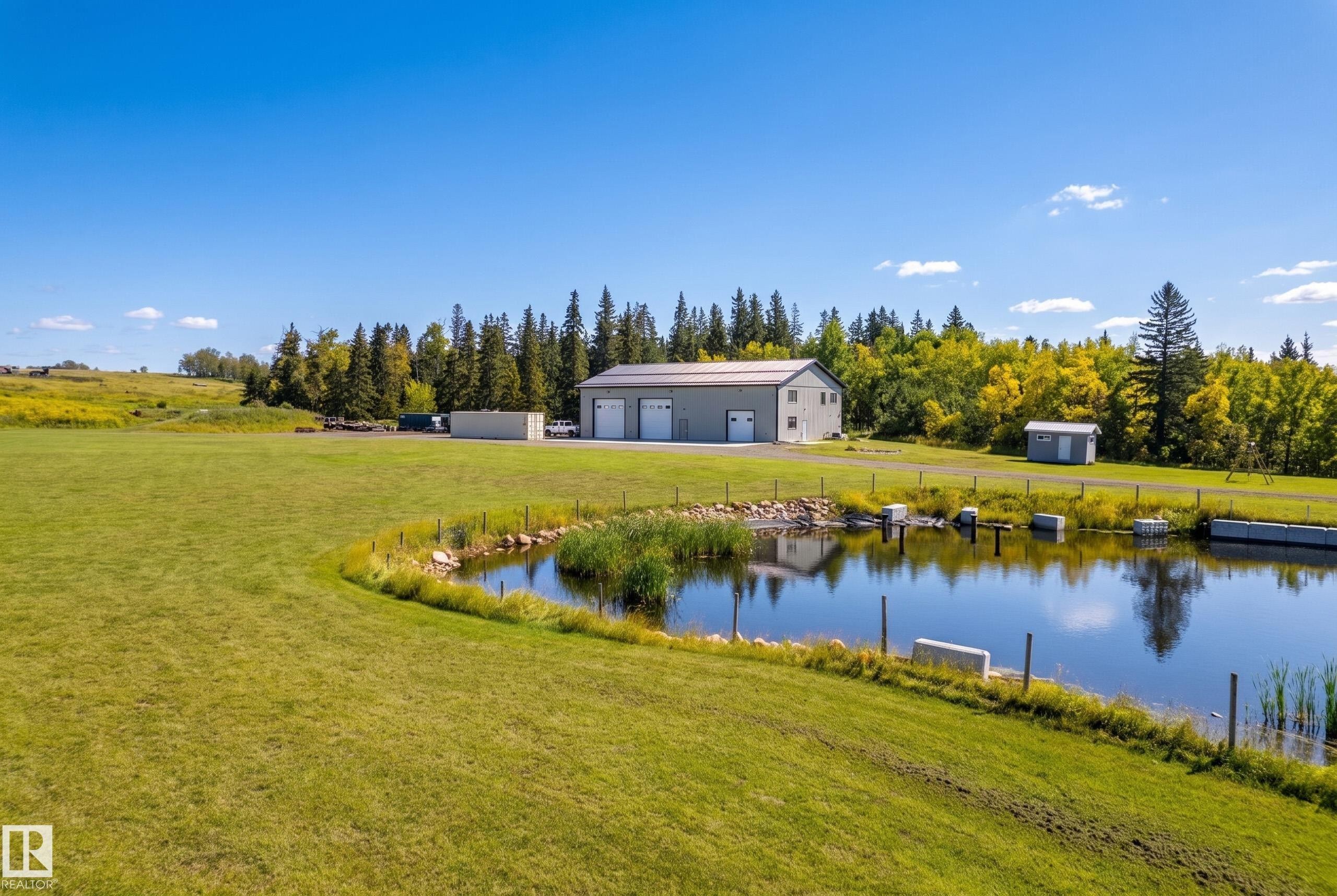 Expansive green space featuring a water body, a large two-story outbuilding with multiple garage doors, and additional storage structures - 49279 Rge Rd 240, Rural Leduc County, AB