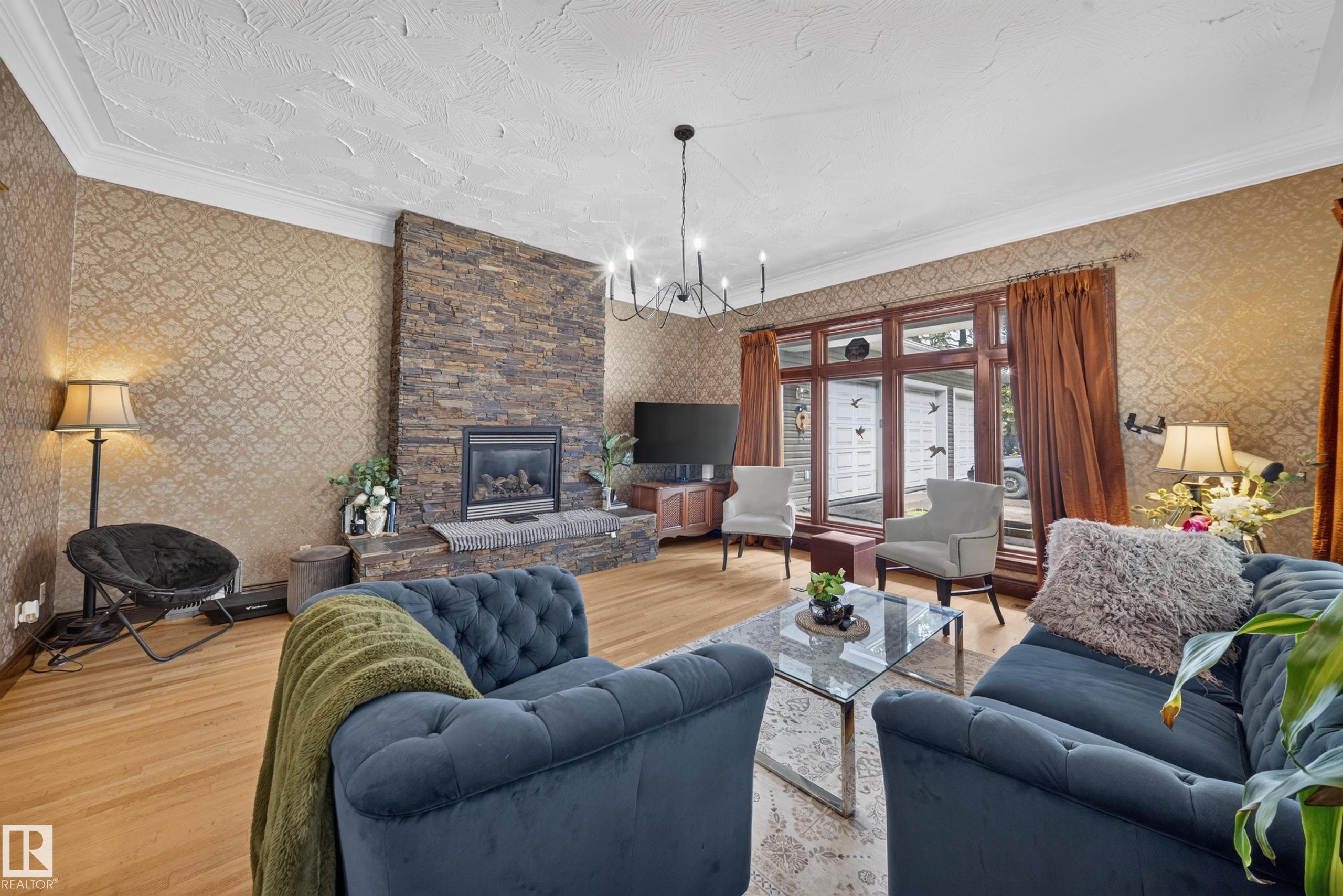 Stacked stone fireplace with a built-in hearth, wood-finish flooring, patterned wall covering, a large window with wood trim, and a chandelier lighting fixture - 719 65 Ave Nw, Edmonton, AB