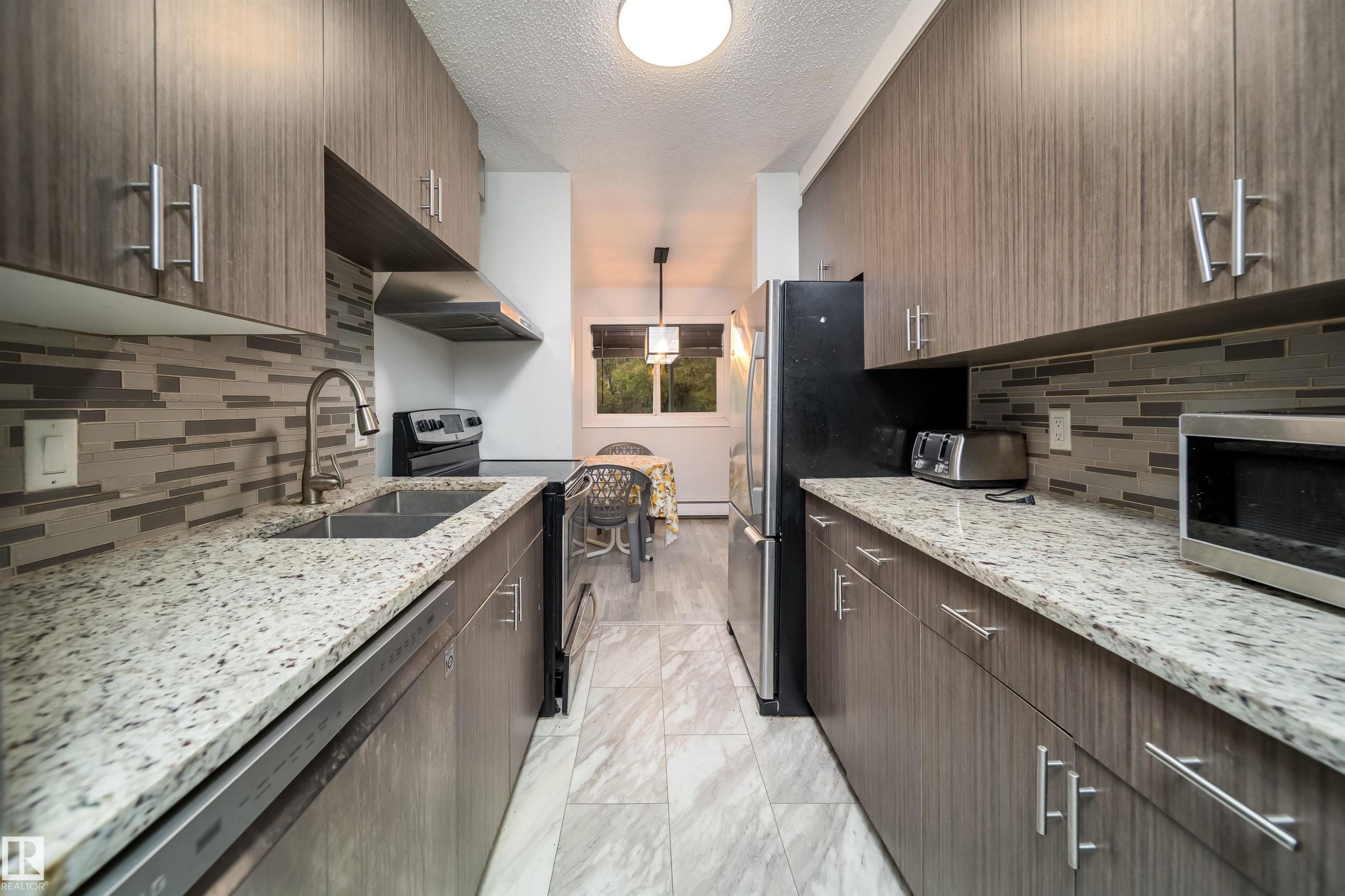 Galley kitchen featuring wood-look cabinetry, granite-style countertops, and a full-height tile backsplash - #202 10615 156 St Nw, Edmonton, AB