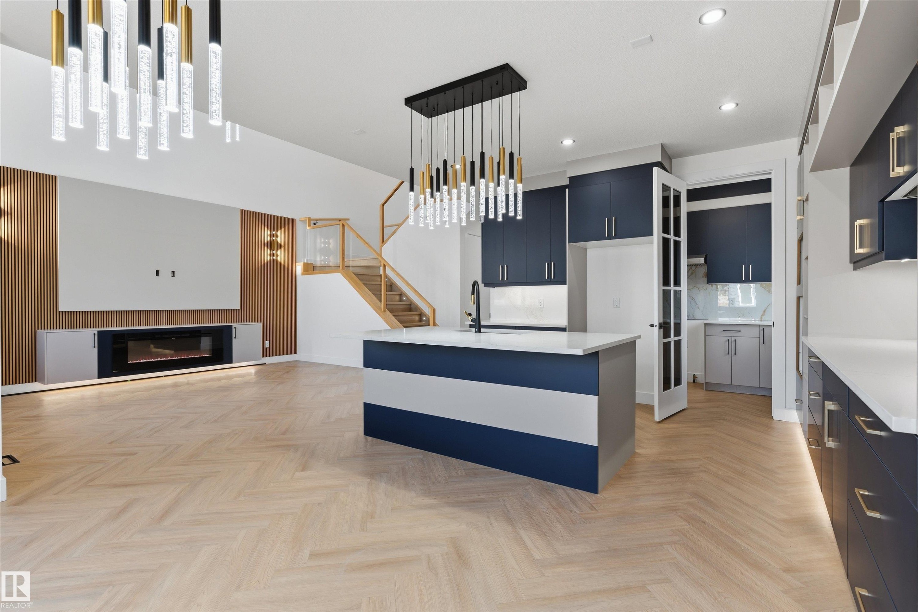 Herringbone wood-finish flooring, two-tone kitchen island with a built-in sink, dark blue cabinetry with gold-tone hardware, contemporary linear fireplace, and a wood-slat accent wall - 2720 63 Avenue, Rural Leduc County, AB