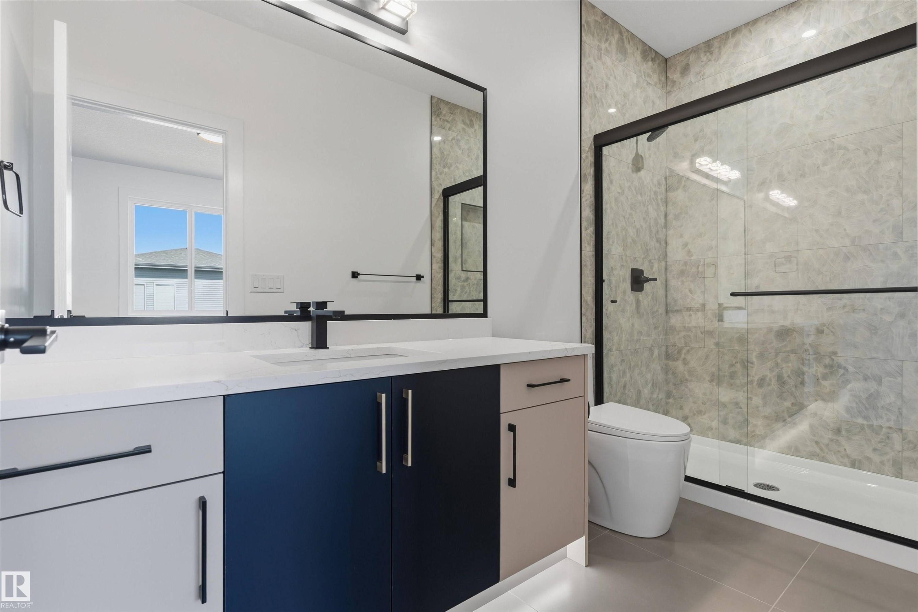 Modern bathroom featuring a vanity with a white countertop and multi-toned cabinetry, an integrated sink, a large framed mirror, and a walk-in shower with a glass enclosure and tiled walls - 2720 63 Avenue, Rural Leduc County, AB