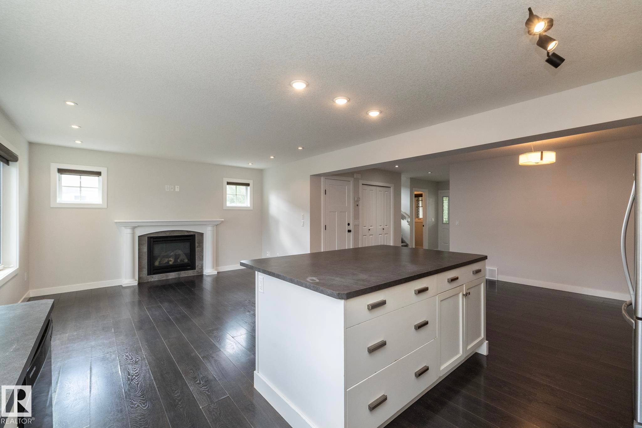Expansive living area featuring dark wood-finish flooring, a central island with white cabinetry, recessed lighting, a fireplace with a white mantel, and a stainless steel refrigerator - 55 Michigan Street, Devon, AB