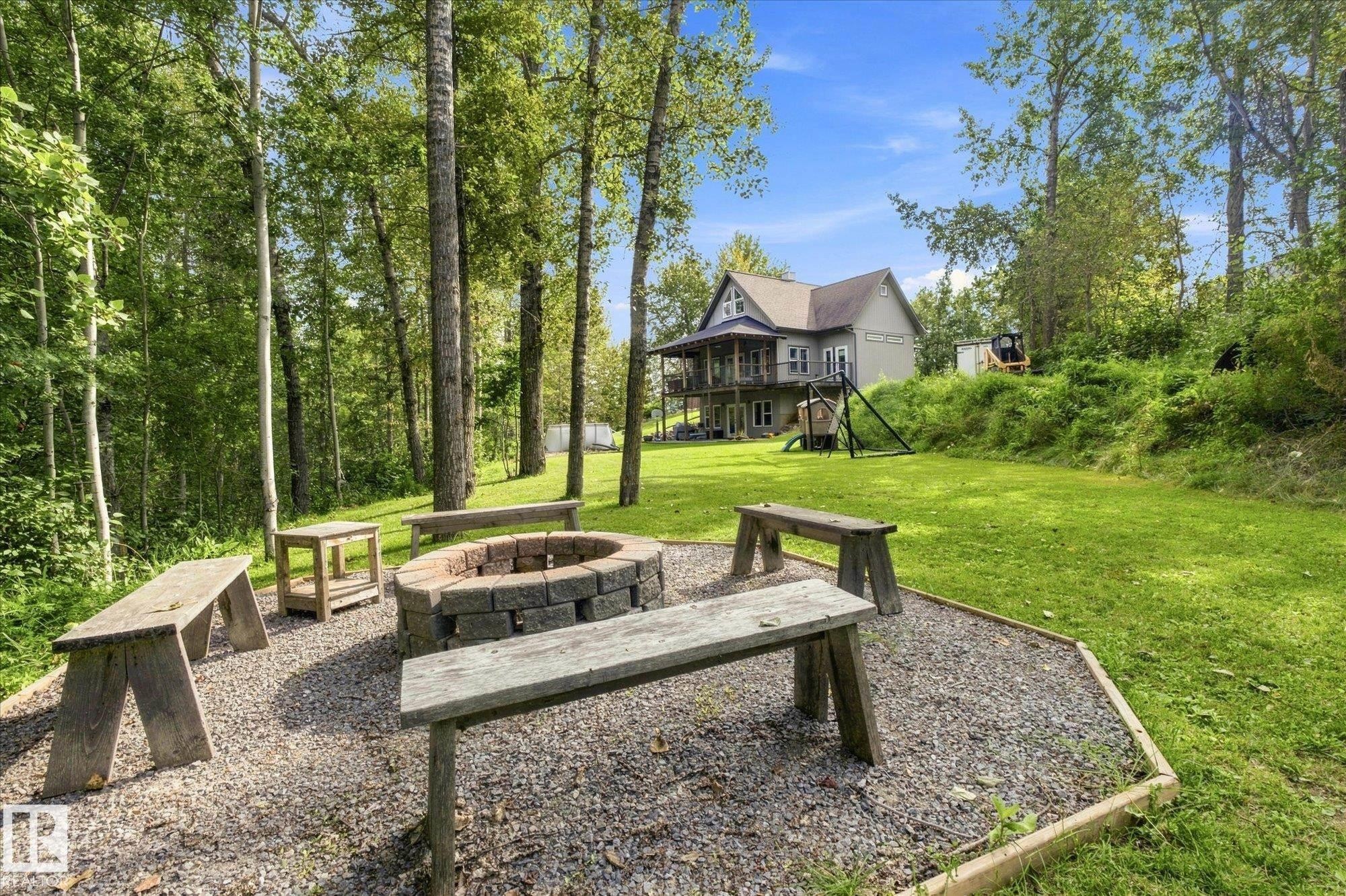 Gravel patio featuring a stone-lined fire pit, surrounded by multiple wooden benches, all set within a landscaped yard with a sloped, treed backdrop - #45 53122 Rge Road 14, Rural Parkland County, AB