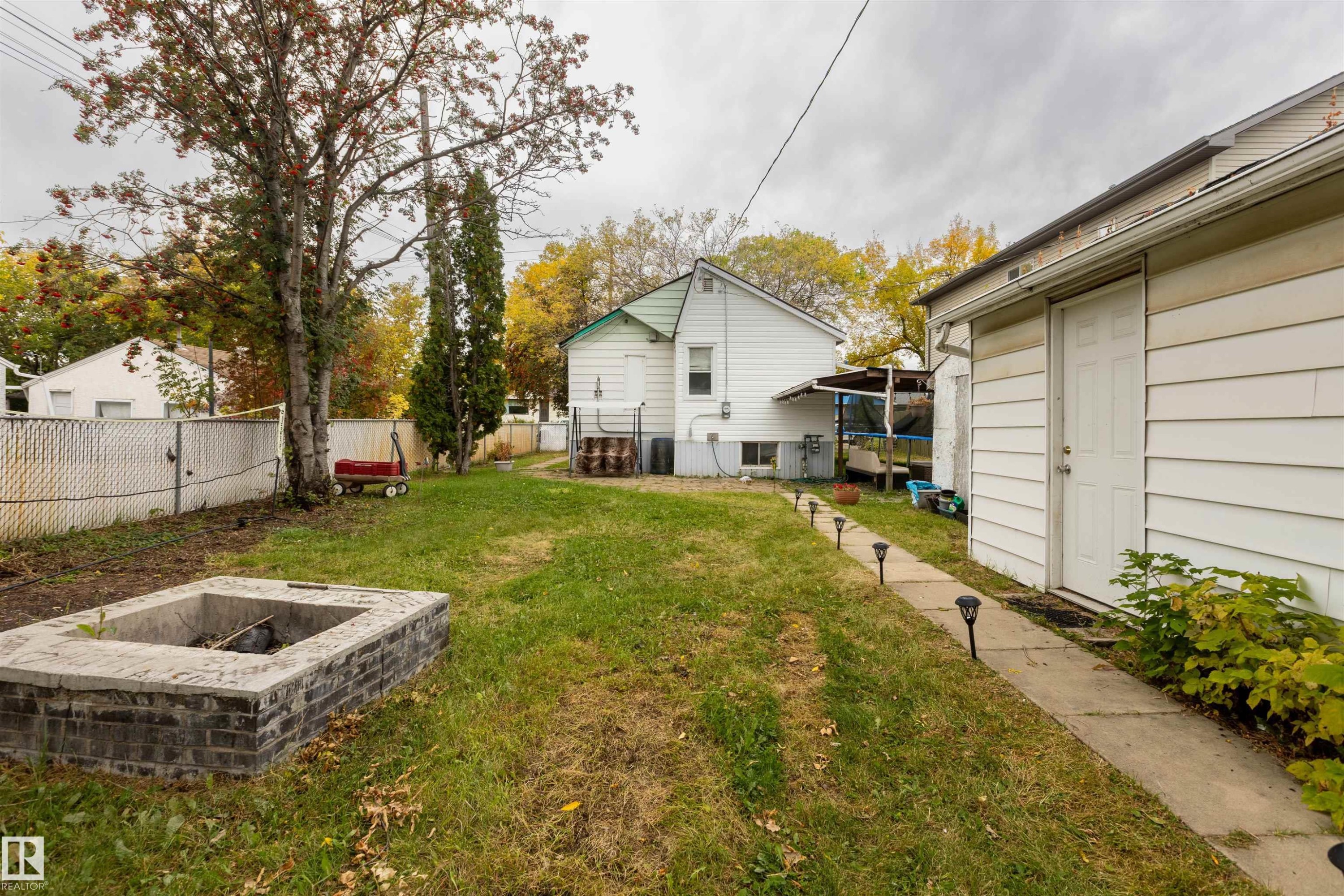 Expansive backyard featuring a built-in fire pit with brick detailing, mature trees, and a concrete pathway with pathway lighting - 12707 73 Street Nw, Edmonton, AB
