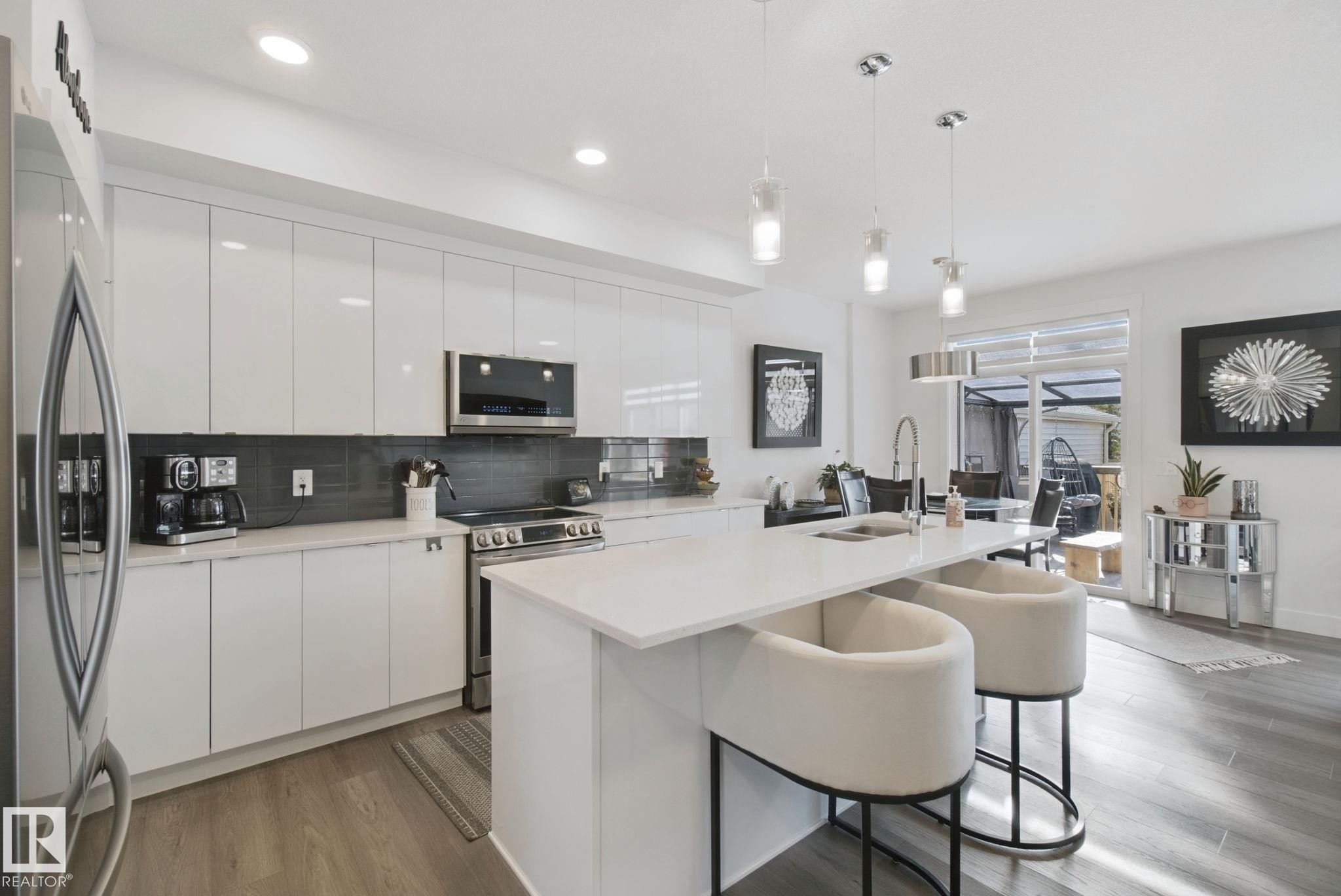 Contemporary kitchen featuring a large island with countertop seating, high-gloss white cabinetry, stainless steel appliances, and a dark tiled backsplash - 153 Cavanagh Common Sw, Edmonton, AB