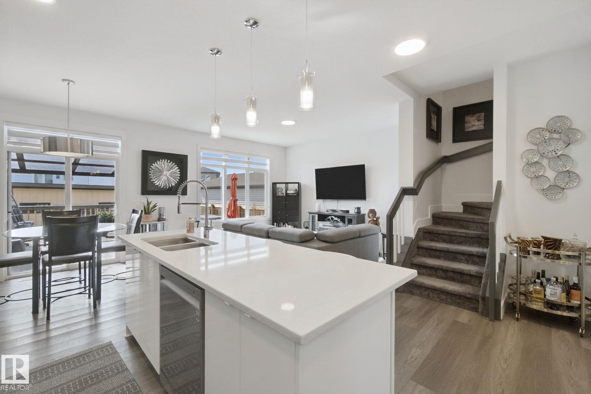 Kitchen island with a white countertop and integrated sink, stainless steel dishwasher, wood-finish flooring, recessed lighting, and a staircase with carpeted treads - 153 Cavanagh Common Sw, Edmonton, AB