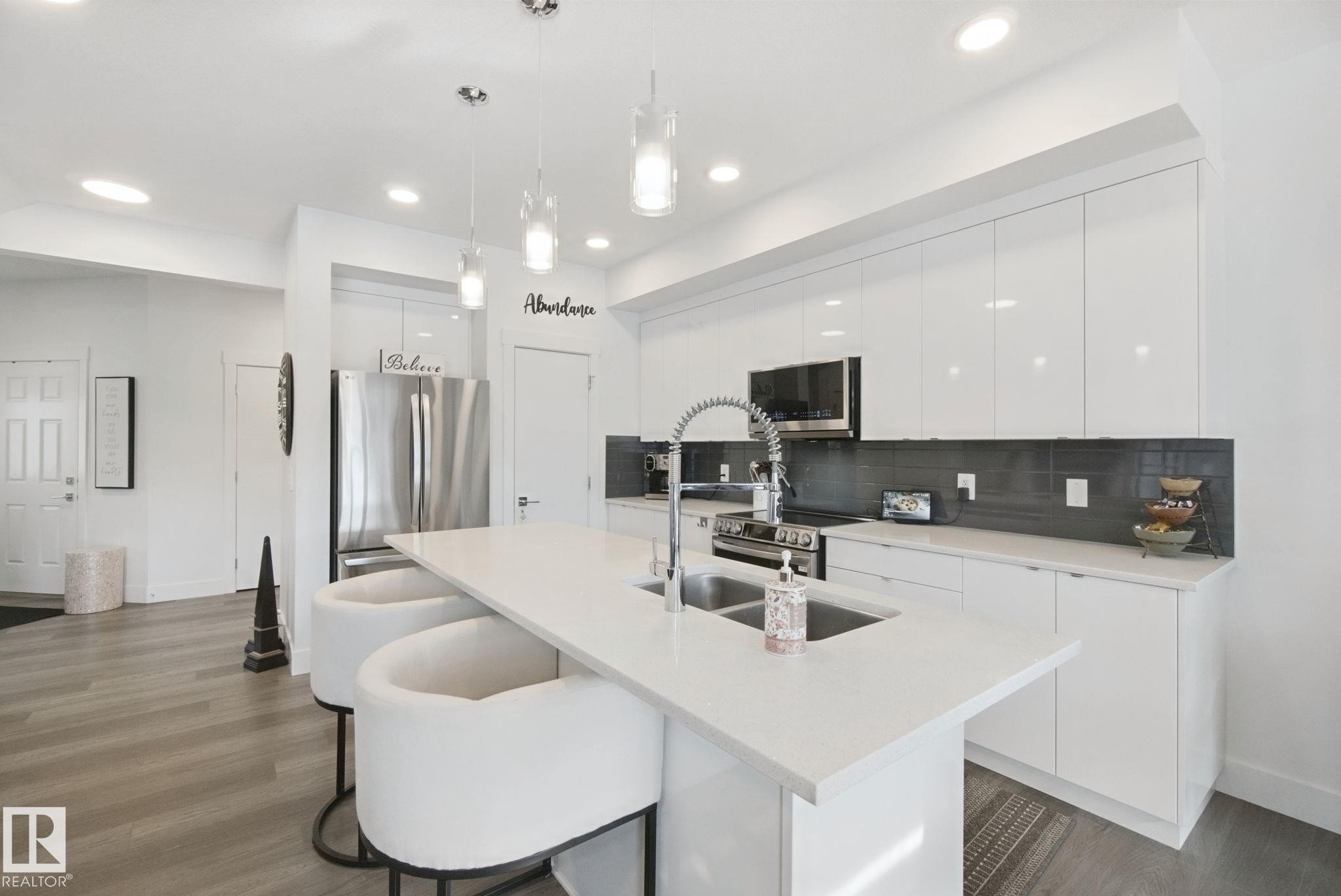 Contemporary kitchen featuring a large center island with an integrated double sink, sleek white cabinetry, stainless steel appliances, and a dark tile backsplash - 153 Cavanagh Common Sw, Edmonton, AB