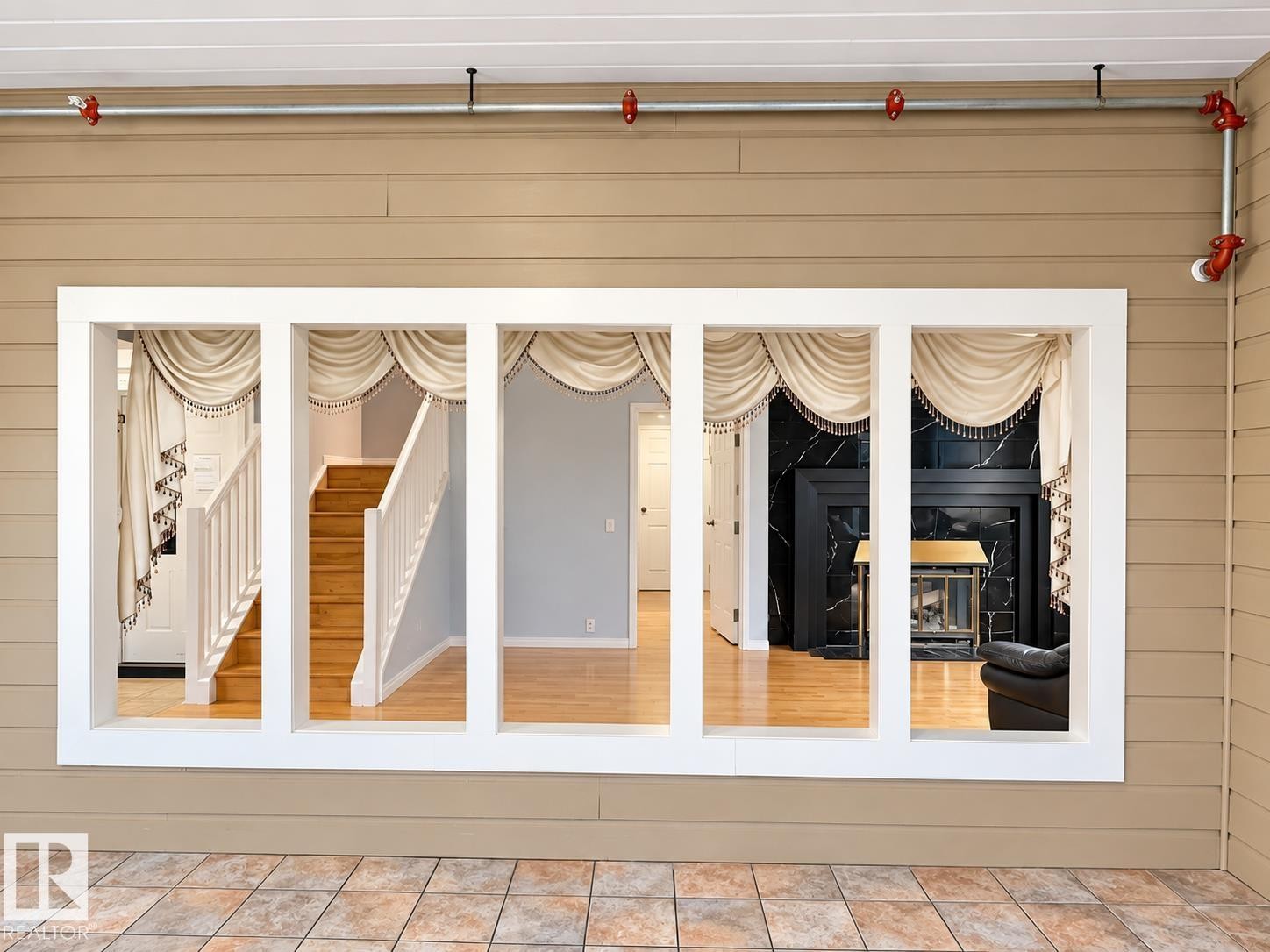 Tiled patio with a multi-pane window looking into an interior with wood-finish flooring, a white-painted staircase, and a fireplace with a black surround - 18929 99A Avenue Nw, Edmonton, AB
