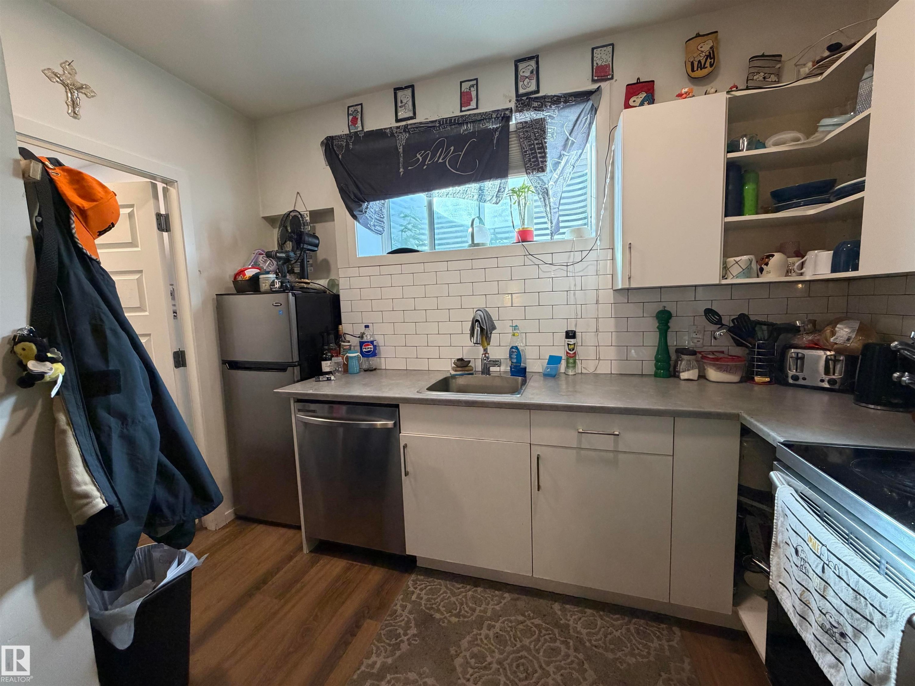 Kitchen featuring wood-finish flooring, white subway tile backsplash, white cabinetry, stainless steel dishwasher, and a window - 11442 82 Street Nw, Edmonton, AB