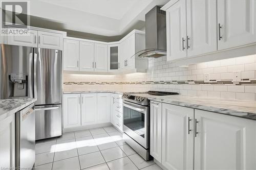 Kitchen with stainless steel appliances, light tile patterned flooring, light stone countertops, wall chimney exhaust hood, and white cabinets - 21 White Star Lane, Grimsby, ON 