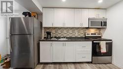 Kitchen featuring light wood finished floors, decorative backsplash, dark countertops, appliances with stainless steel finishes, and white cabinetry - 