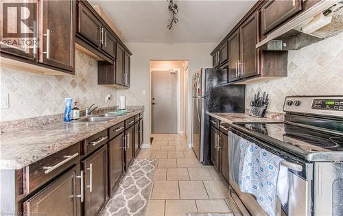 Kitchen featuring stainless steel electric range oven, under cabinet range hood, dark brown cabinets, decorative backsplash, and light tile patterned floors - 283 Fairway Road N Unit# 501, Kitchener, ON - Indoor Photo Showing Kitchen With Double Sink