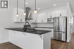 Kitchen featuring appliances with stainless steel finishes, hanging light fixtures, a peninsula, white cabinetry, and wall chimney range hood - 