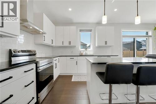 Kitchen with stainless steel electric stove, a kitchen breakfast bar, wall chimney range hood, decorative light fixtures, and dark wood-style flooring - 7266 Silver Creek Circle, London, ON - Indoor Photo Showing Kitchen With Upgraded Kitchen