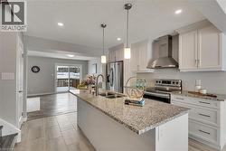 Kitchen featuring wall chimney exhaust hood, a center island with sink, stainless steel appliances, hanging light fixtures, and light stone countertops - 