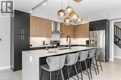 Kitchen featuring a kitchen breakfast bar, stainless steel appliances, decorative light fixtures, wall chimney range hood, and a center island with sink - 