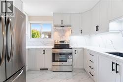 Kitchen featuring appliances with stainless steel finishes, light marble finish flooring, light stone countertops, white cabinetry, and under cabinet range hood - 