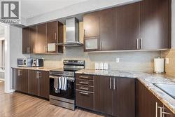 Kitchen with appliances with stainless steel finishes, dark brown cabinetry, wall chimney exhaust hood, light wood-style floors, and light stone countertops - 