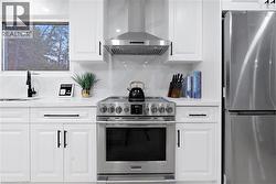 Kitchen featuring appliances with stainless steel finishes, wall chimney exhaust hood, white cabinetry, light stone countertops, and backsplash - 