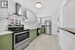 Kitchen featuring green cabinets, stainless steel appliances, white cabinetry, wall chimney range hood, and light stone counters - 