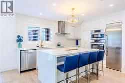 Kitchen with a breakfast bar area, white cabinetry, stainless steel appliances, wall chimney exhaust hood, and light stone counters - 