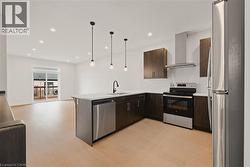 Kitchen featuring stainless steel appliances, wall chimney exhaust hood, dark brown cabinetry, light hardwood flooring, and a peninsula - 