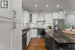 Kitchen featuring glass insert cabinets, stainless steel appliances, white cabinetry, wall chimney range hood, and dark stone counters - 